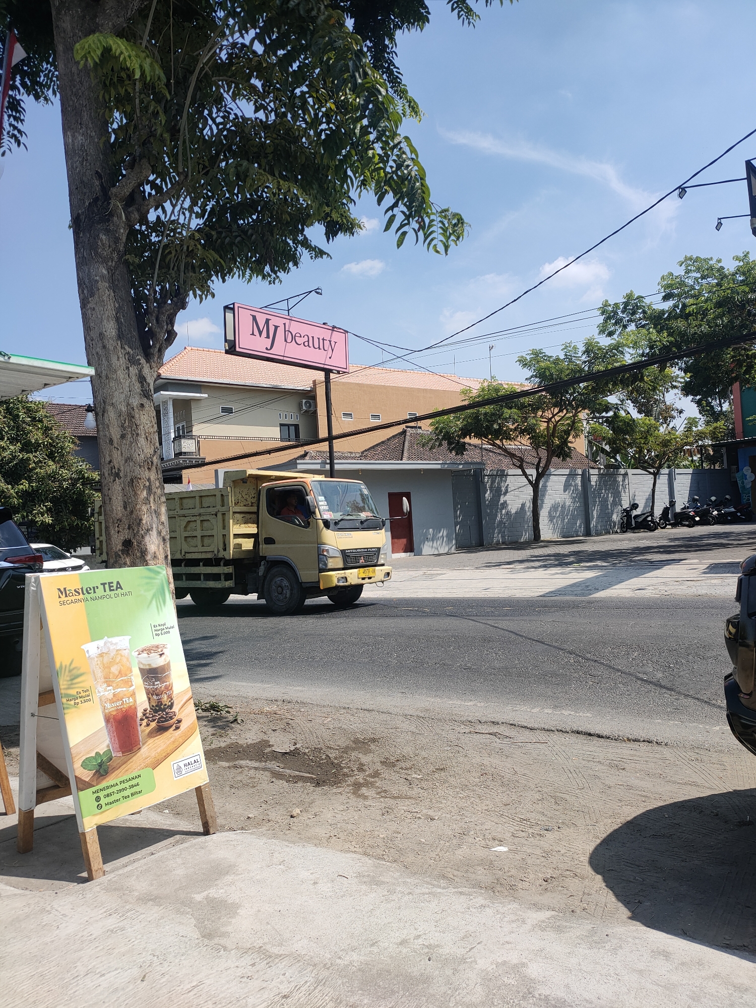 In Kanigoro, Indonesia, on a bright, sunny day, a yellow dump truck is parked on the side of a paved road. Adjacent to the truck, a tall tree with green foliage stands to the left of the frame. In the foreground, a freestanding sign displays promotional images of drinks and text, with "Master TEA" prominently featured. Behind the truck, a building with a tiled roof and light-colored walls is visible. Further in the background, more trees line a grey brick wall, and several motorcycles are parked. The sky above is clear blue with a few wispy clouds, and power lines crisscross the scene.