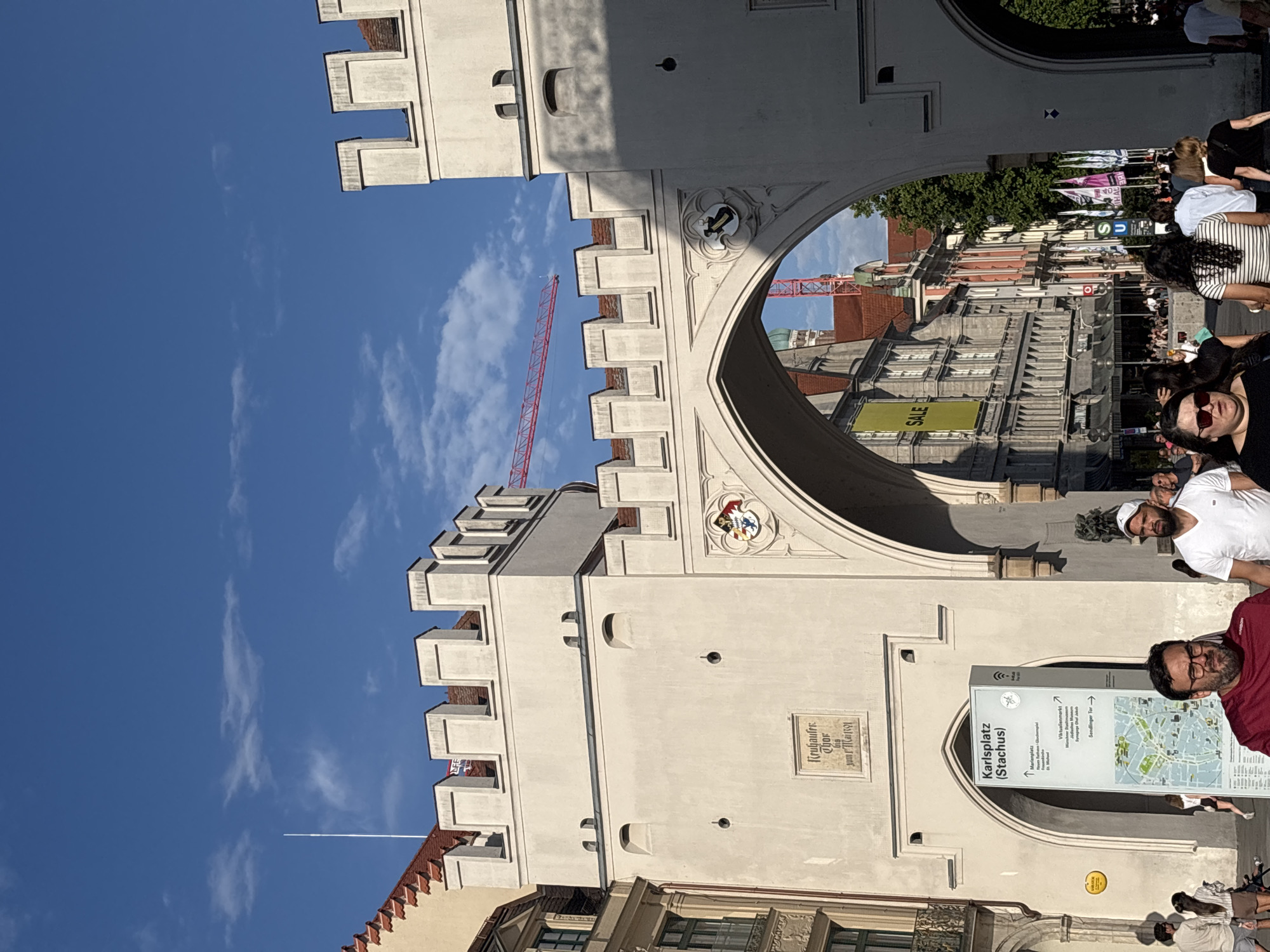 The image captures a view through the Karlstor, also known as Stachus, a historic gate in Munich, Germany. The gate's pale stone structure with crenellated battlements and arched passageways dominates the foreground. A signpost for "Karlsplatz (Stachus)" stands to the left of the central archway. Beyond the arch, a bustling street scene unfolds with people walking, shops with awnings, and flags displayed. The architecture of the buildings lining the street is varied, with some featuring classical elements and others more modern facades. A bright pink construction crane is visible in the distance, silhouetted against a clear blue sky with scattered clouds. The light suggests it is daytime, likely late afternoon given the shadows. Several individuals are in focus in the foreground, including a man in a red shirt and glasses and another in a white t-shirt and white cap, walking towards the viewer. Other pedestrians are seen in the background, some appearing to be tourists based on their attire and the presence of a stroller. The overall impression is of a lively urban environment at a significant historical landmark.