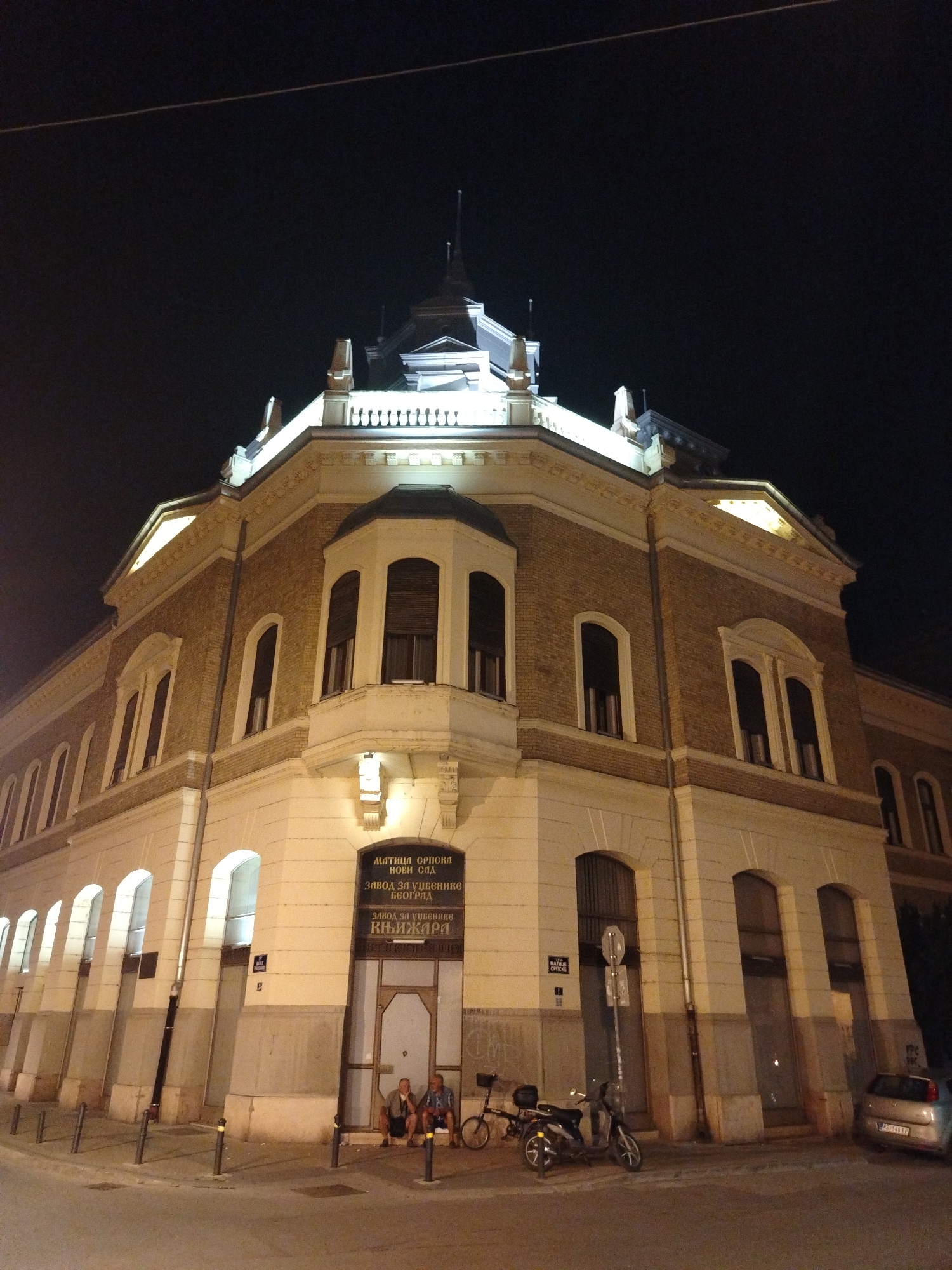 The image shows a corner of a multi-story building at night in Novi Sad, Serbia. The building is constructed of light-colored brick with white decorative elements and trim. It has several arched windows and a bay window on the corner. The building is illuminated by artificial lights.
Above the ground floor entrance, there is a sign with Cyrillic text. The visible text reads "МАТИЦА СРПСКА НОВИ САД" and "ЗАВОД ЗА УЏБЕНИКЕ БЕОГРАД" and "КЊИЖАРИ". This indicates that the building houses the Matica Srpska in Novi Sad, and the Institute for Textbooks in Belgrade, as well as a bookstore.
On the ground floor, two men are sitting on the steps in front of the entrance. A bicycle and a scooter are parked nearby on the sidewalk. A silver car is parked on the street to the right of the building. The street is paved and there are streetlights and bollards visible. The sky is dark, indicating it is nighttime.