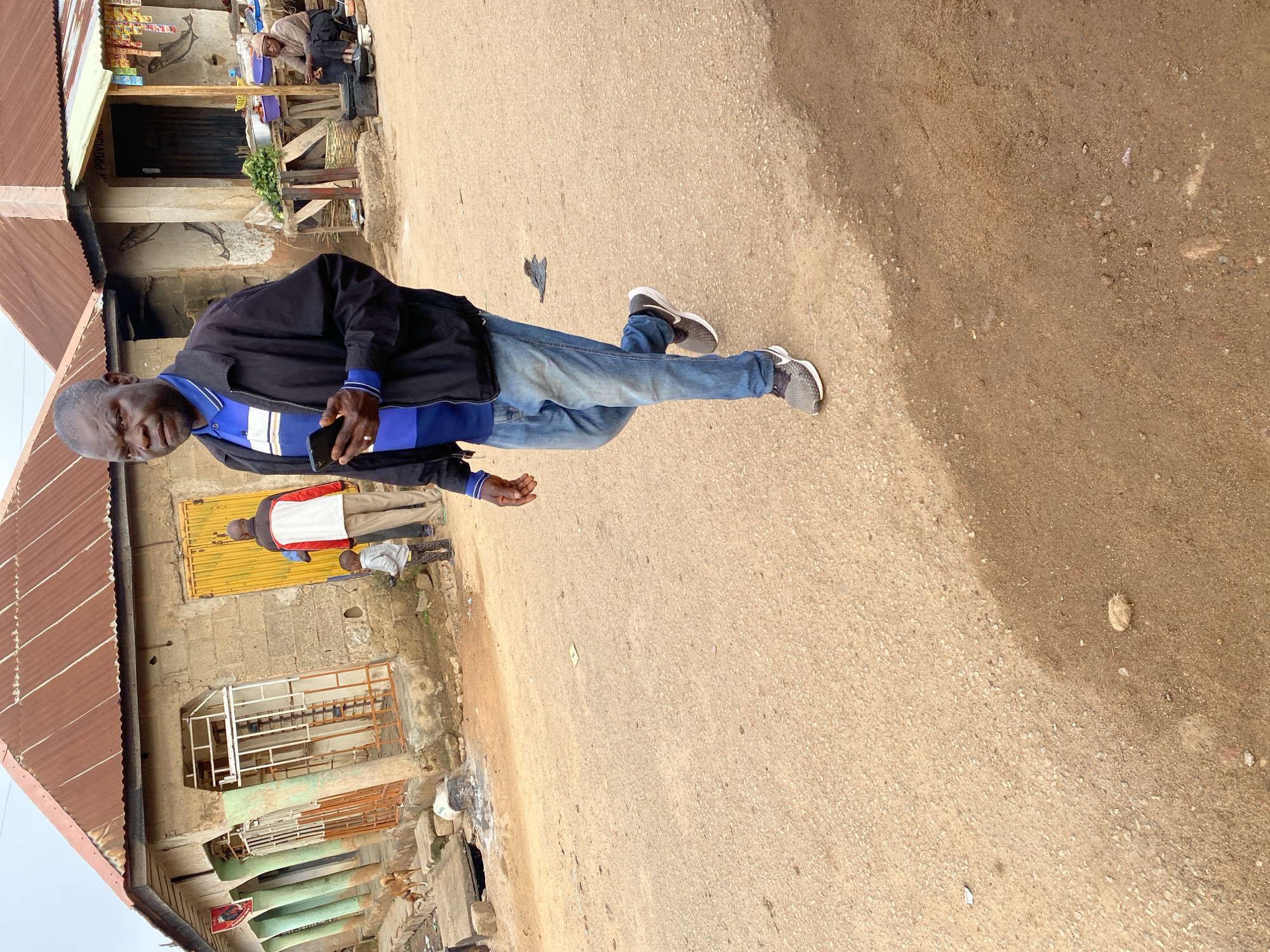 In Jos, Nigeria, a man in a dark jacket and blue jeans walks on a dirt road in front of a building with a rusty corrugated metal roof. He holds a smartphone in his right hand. In the background, a child stands next to a man in a red and white jacket. A dog is visible to the left of the building, and another person sits on a stool near a stall selling produce. The area appears to be a market or a commercial street, with various stalls and small shops lining the road. The overall environment is dusty and appears to be an outdoor setting in a developing urban area.
