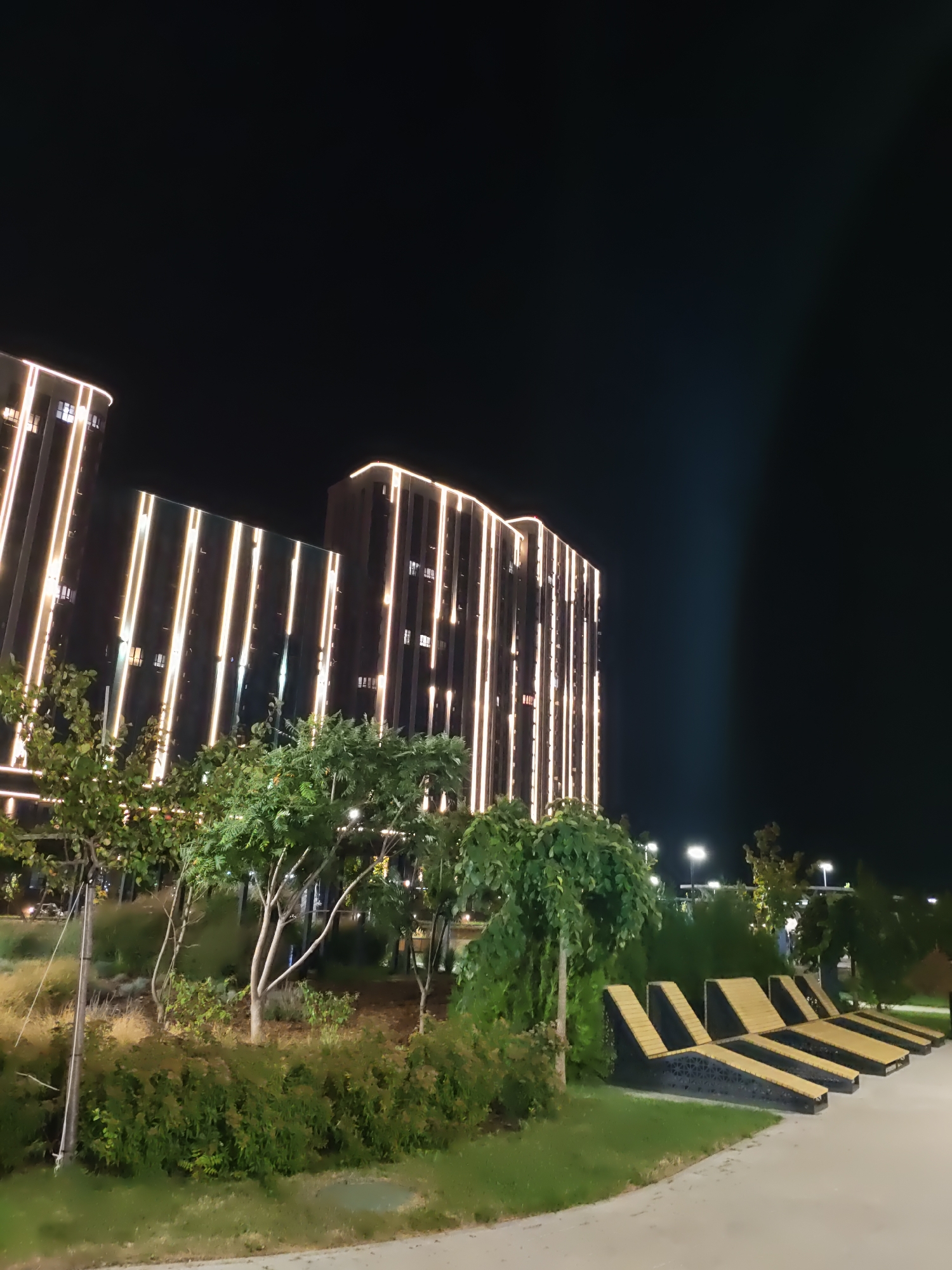 This image depicts a nighttime scene in the city of Rostov-on-Don, Russia, likely in a park or public space. In the background, two modern, multi-story buildings are illuminated by vertical lines of warm white lights that trace their facades. The buildings appear to be residential or office complexes. In the foreground, there is a landscaped area with green bushes, trees, and a grassy slope. Several wooden lounge chairs are arranged in a row on a paved surface, suggesting an area for relaxation or outdoor seating. The sky is dark, indicating it is nighttime, with a faint light beam visible in the upper right quadrant of the image. No people are visible in the scene.