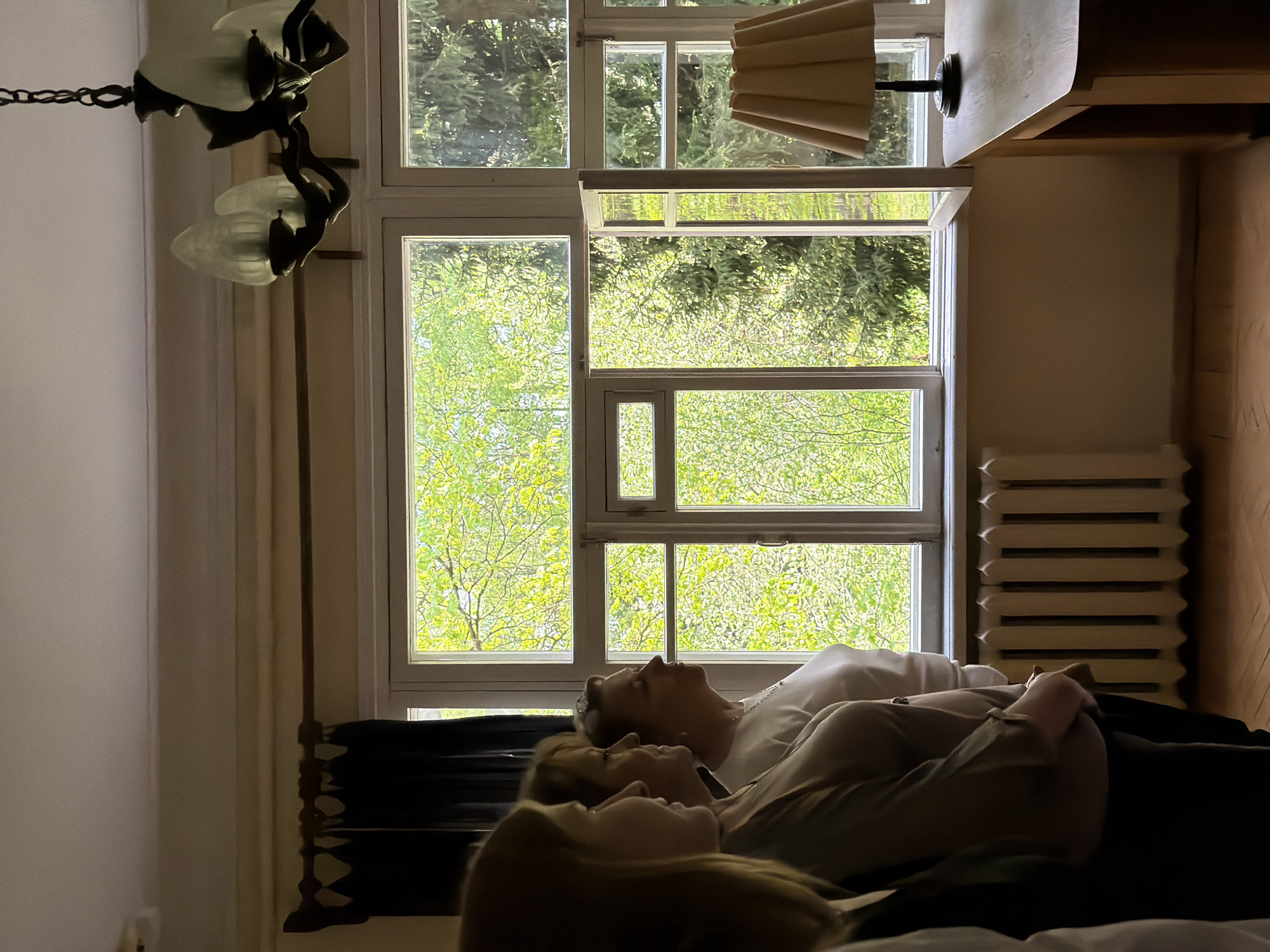 Three people are lying down in a room. A large window dominates the back wall, offering a view of green trees, likely outside. A radiator sits to the right of the window. A chandelier hangs from the ceiling on the left side of the image. A desk lamp is above the window. Based on the architecture, this image was most likely taken in Russia. Although Odintsovo is a possibility, it is difficult to verify the precise location using the provided information.