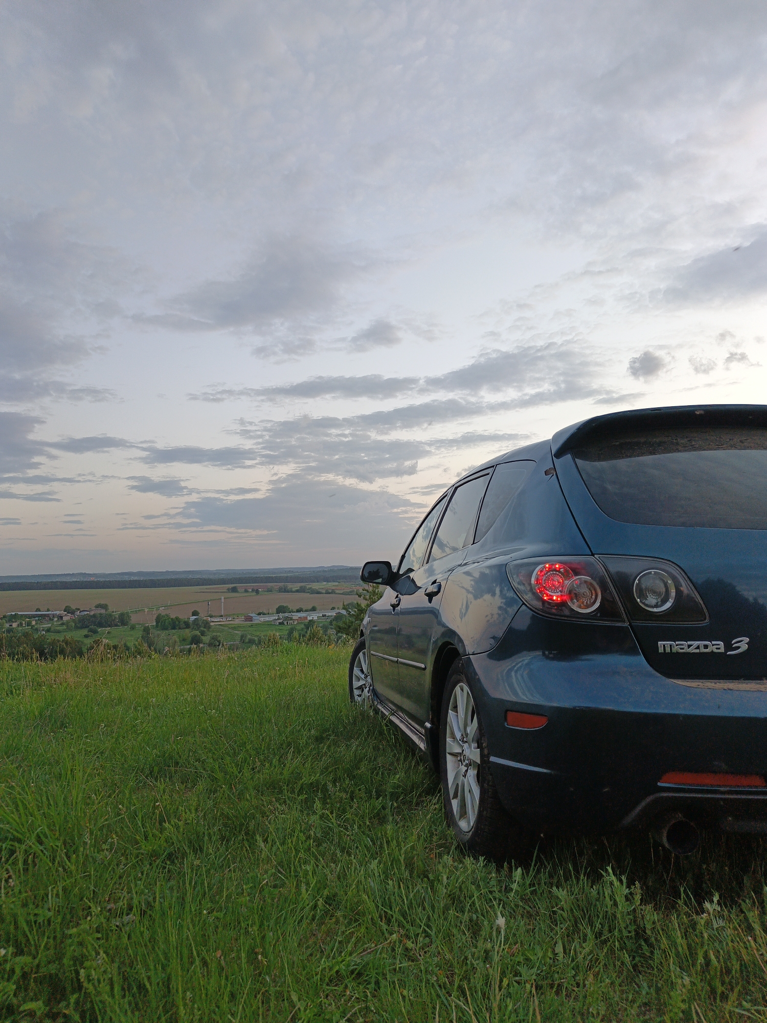 The image shows a dark blue Mazda 3 hatchback parked on a grassy hill. The rear of the car is prominently featured, with the "mazda 3" logo visible. The brake light is illuminated on the left side. The car is parked in an area of tall green grass. Beyond the grass, there is a view of a rural landscape with fields and trees. The sky is cloudy and suggests dusk or dawn. The location is in a field of grass in Чумна, Russia.