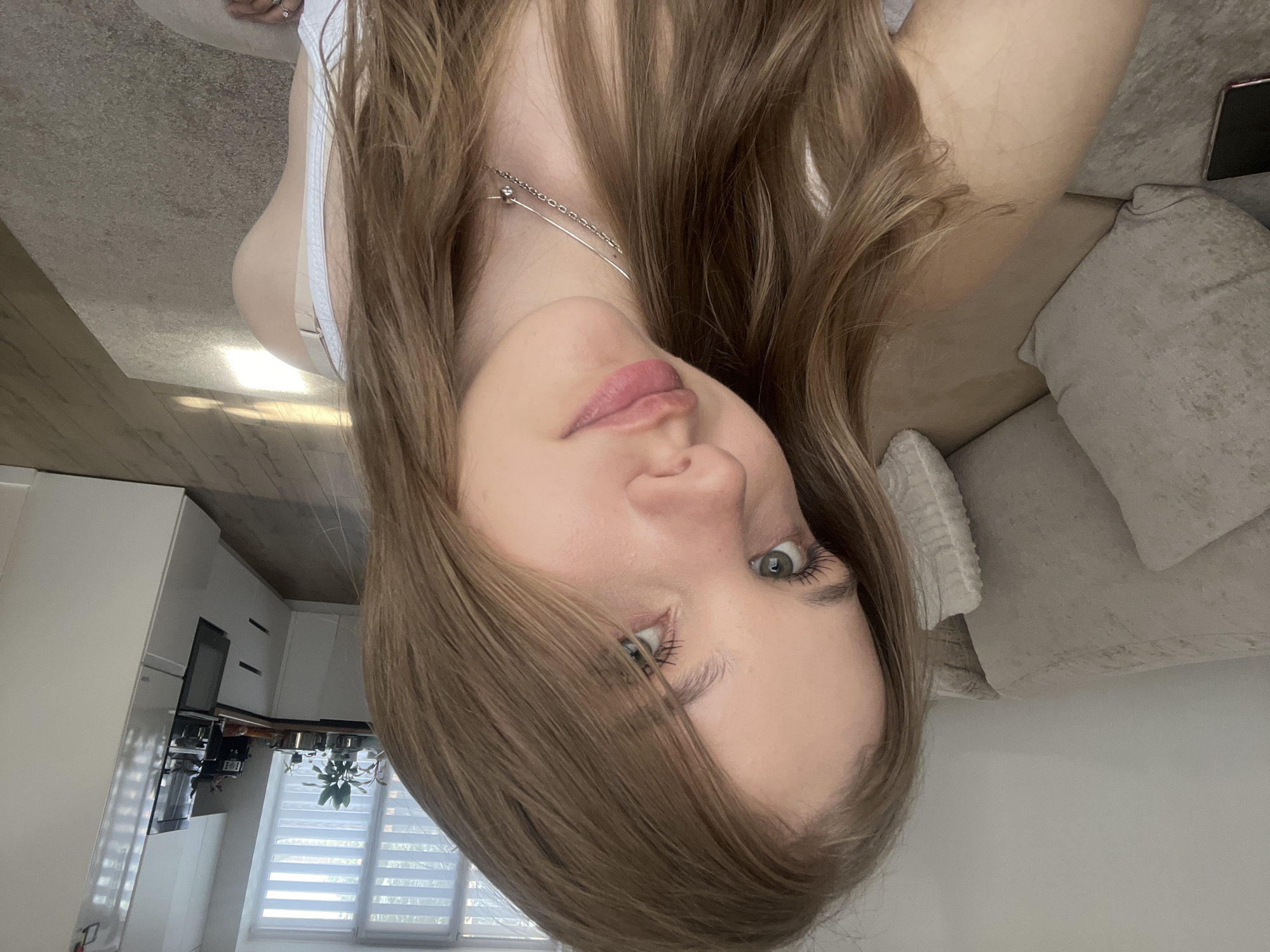 A young woman with long brown hair is the primary subject. She is wearing a light-colored top and a silver necklace. She is facing the camera with a slight tilt. Her eyes are light-colored.
The background includes a kitchen with white cabinets and appliances visible through a window with blinds. Part of a living room is visible, featuring a beige sofa and carpet.
The location in Votkinsk, Russia is not discernible from the image. There are no specific markers, landmarks, or elements in the image that can definitively identify the location as Votkinsk.
