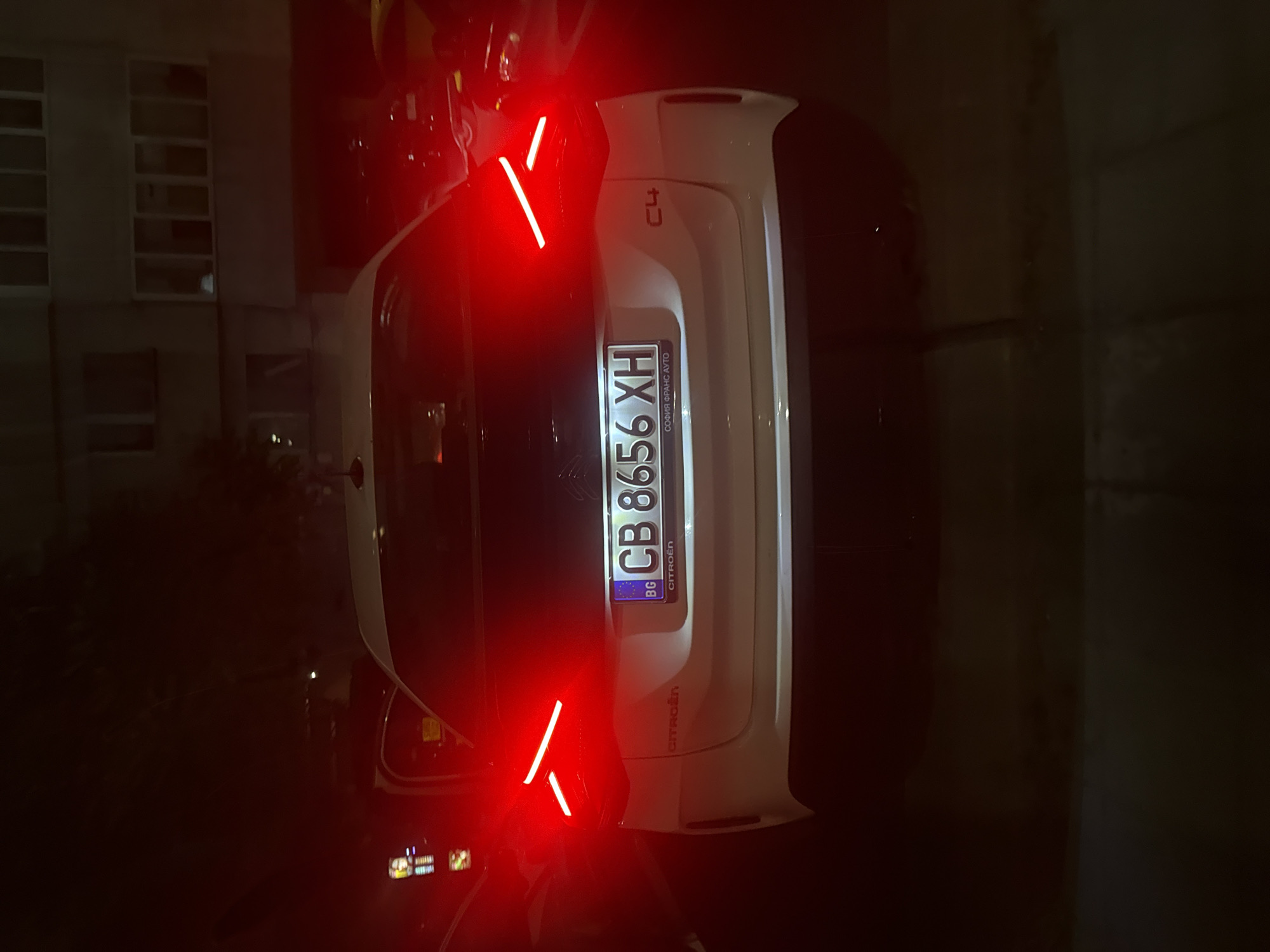 A white Citroen C4 hatchback is parked at night, its rear lights illuminated red. The car is facing away from the camera, with its license plate clearly visible: CB 8656 XH. The license plate also has a blue European Union strip on the left and the Bulgarian abbreviation "BG". Below the license plate, the text "CITROËN" is visible, and to the right, "C4". The car is parked in front of a residential building with balconies and windows. The scene appears to be in an urban environment, likely a parking area or street in the city of Burgas, Bulgaria, given the Bulgarian license plate and the style of the building. No people are visible in the image.