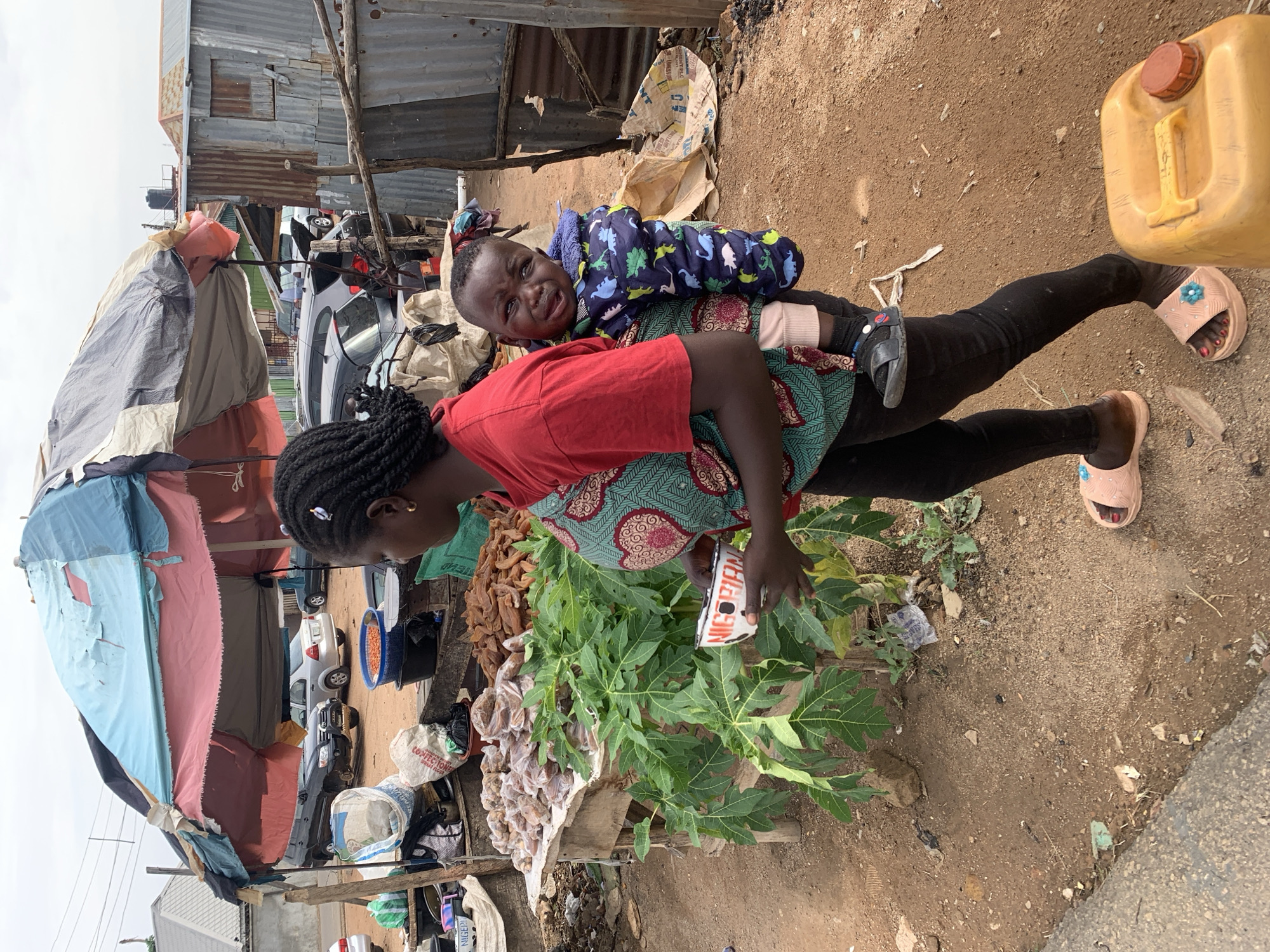 An outdoor scene in Jos, Nigeria, captures a woman with a young child strapped to her back, attending a street market stall. The woman wears a red top, a patterned green and red wrapper, black leggings, and light pink slides with a blue flower detail. Her braided hair is visible, and she looks down and to her left, her face partially obscured. The child, a toddler, is dressed in a blue puffer jacket with dinosaur patterns and black shoes, looking towards the camera with a mixed expression that could be a slight grimace or a nascent smile.
In front of the woman is a makeshift stall laden with various items. Large green leafy branches, possibly papaya leaves, dominate the foreground. Beside them are piles of dried brown produce and smaller items neatly packaged in clear plastic bags. A white bag, partially visible, displays the word "CONFECTION...". The woman holds a small red and white container, likely a cup or bowl, with "NIGERIAN" clearly visible on its side.
The ground is unpaved, consisting of dirt and scattered debris. To the right of the woman, a large yellow plastic jerry can with a red cap rests on the ground. The background reveals an informal settlement or market area, characterized by several structures constructed from corrugated metal sheets, some appearing multi-storied. Cars are parked further back, and a water tank is visible on a distant rooftop. To the far left, a stall covered by a blue and red tarp suggests a bustling market environment. The sky is overcast, indicating daytime.