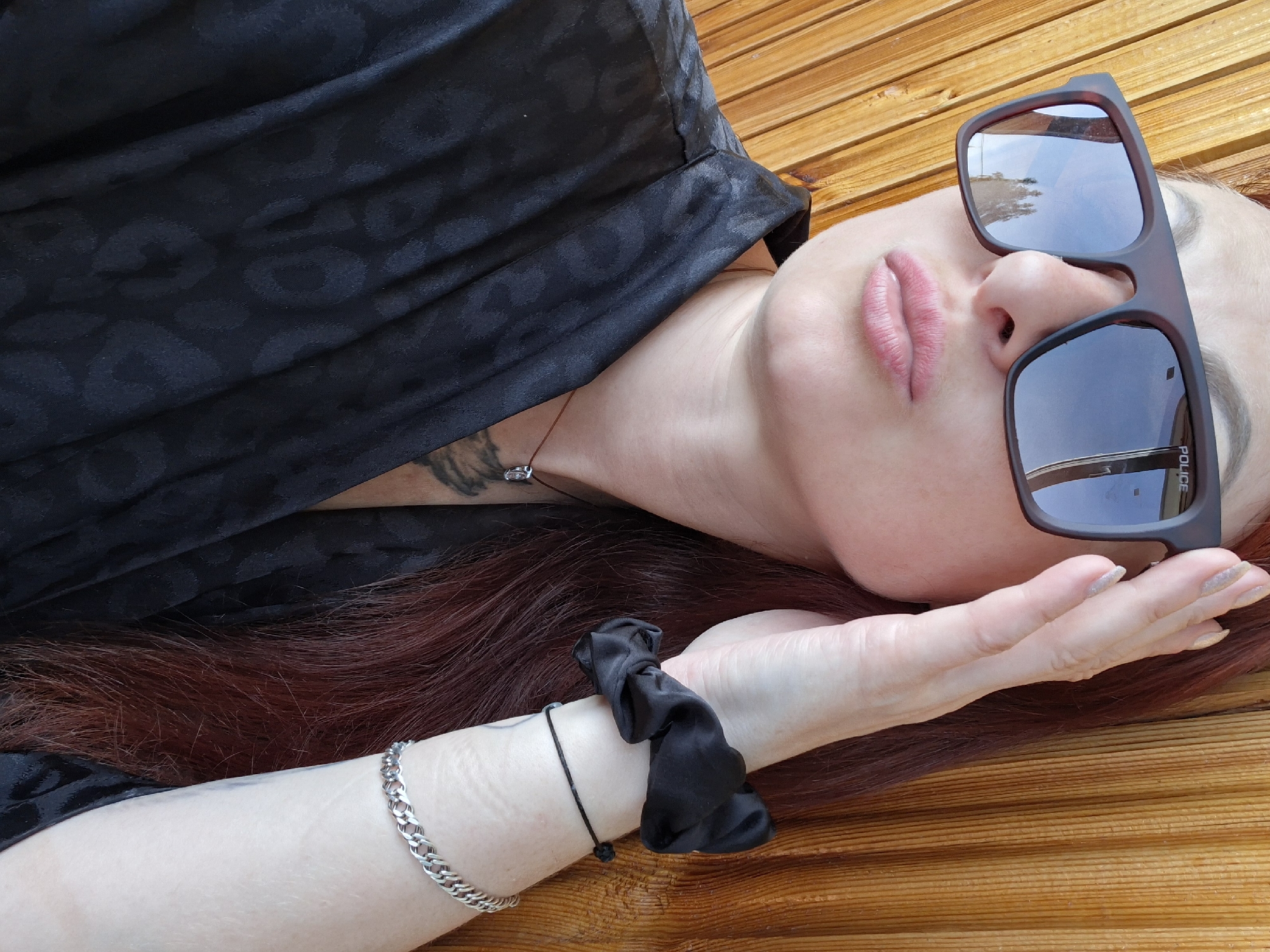 A woman lies on a wooden surface, wearing sunglasses and a black top with a subtle leopard print. Her eyes are covered by the dark lenses of her sunglasses, but her pink lips are slightly parted. Her long, auburn hair spreads out around her. On her wrist, she wears a silver chain bracelet and a black scrunchie. A thin necklace with a small, clear pendant is visible around her neck. The lighting suggests it is daytime. The wooden surface appears to be part of an outdoor bench or seating area. The reflection in her sunglasses shows a cloudy sky and some greenery. Text on the sunglasses reads "POLICE".