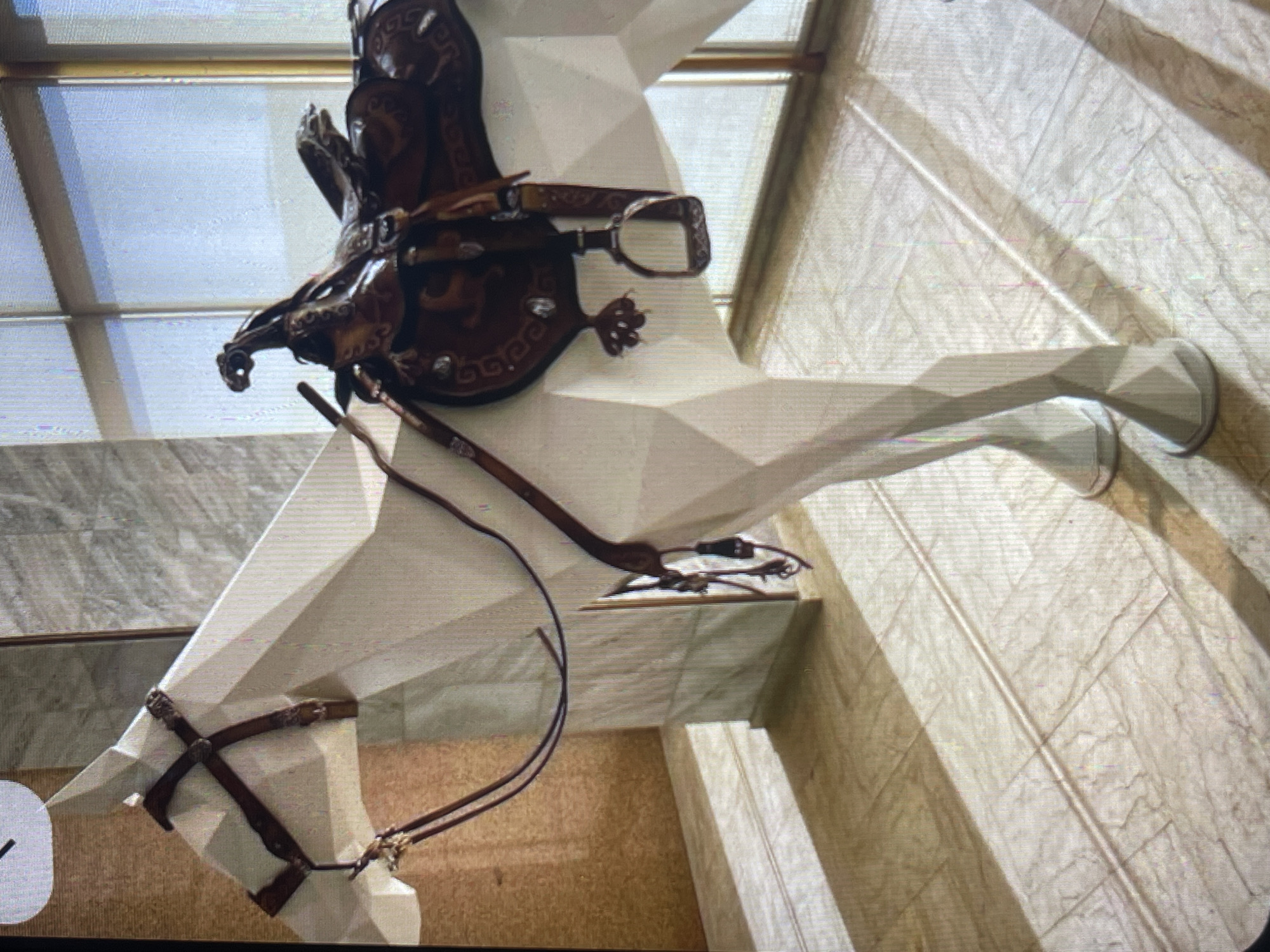 A white, angular, abstract sculpture of a horse is adorned with a dark brown saddle and bridle, positioned indoors on a tiled floor. The horse's form is constructed from geometric planes. Sunlight streams in from the right, casting distinct shadows across the floor and the sculpture. The background features vertical blinds or windows and textured wall panels. The overall impression is of an art installation within an interior space. The location in Bishkek City, Kyrgyzstan, cannot be determined from the visual information provided.