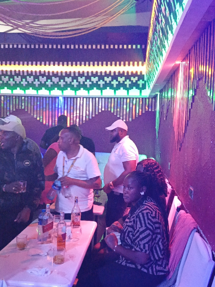 Several people gather at a table in a dimly lit bar or club. A woman sits at the edge of the table in a patterned shirt. Bottles of alcohol and glasses rest on the white surface. Overhead, intricate lighting designs adorn the ceiling and walls, featuring geometric patterns and vibrant colors like green, white, and shades of purple. Other people stand around the table, engaged in conversation.