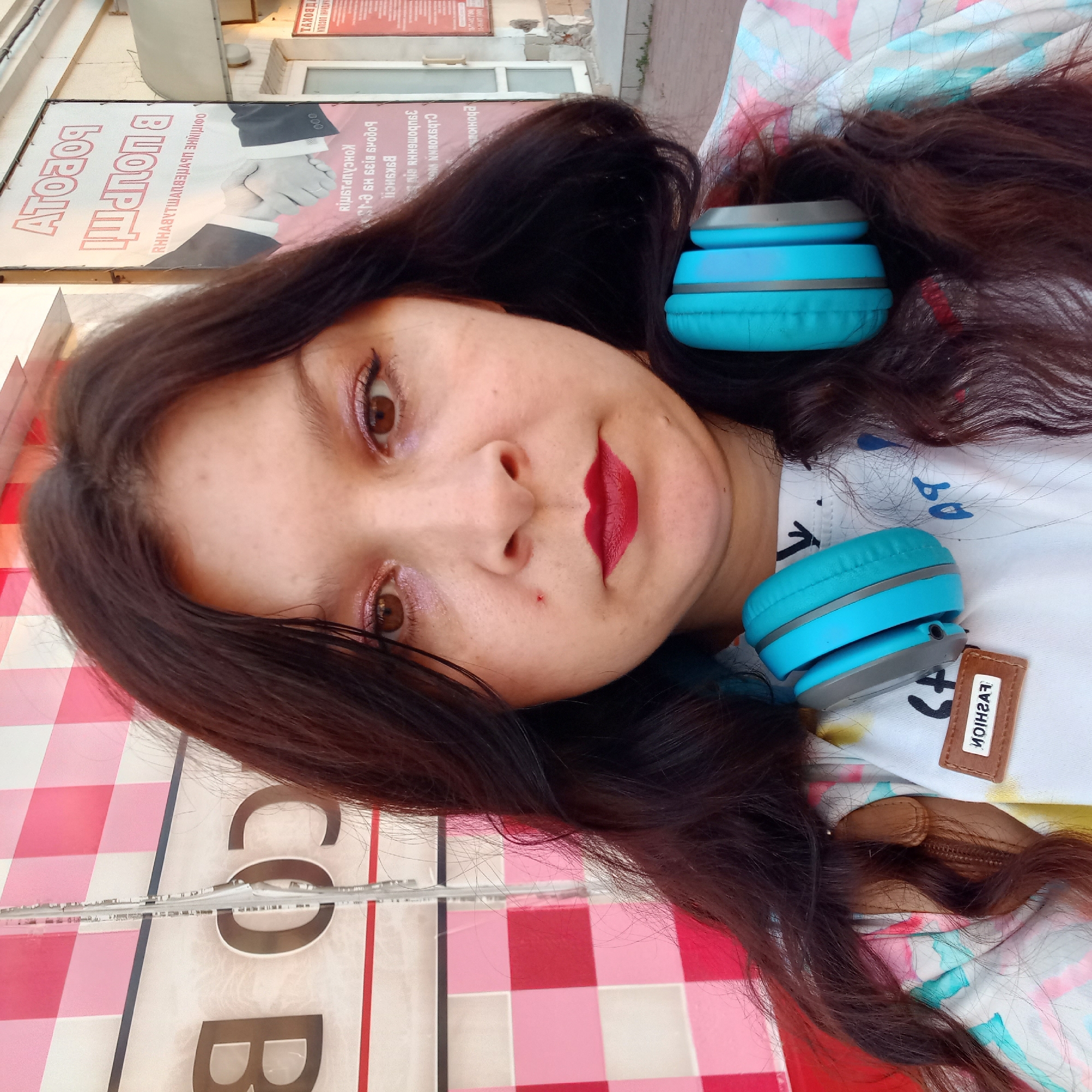 A young woman with dark, wavy hair is taking a selfie outdoors. She is wearing headphones around her neck, which are bright blue with gray accents. She has on a white t-shirt with a tie-dye pattern of pink, blue, and green. On the t-shirt, there is a small brown tag with the word "FASHION" printed in white letters. Her face is centered in the frame, and she is looking directly at the camera. She has brown eyes, and her makeup includes pink eyeshadow and red lipstick. A small red mark is visible on her cheek, just below her left eye.
The background consists of a store facade. To the left of the woman, there is a wall with a pink and white checkered pattern. To her right, a large sign with Cyrillic text in red and white is visible. The text on the sign appears to be an advertisement. Based on the Cyrillic text, it is highly probable that this image was taken in Kropyvnytskyi, Ukraine. The overall environment suggests an urban setting, possibly a commercial street. The lighting indicates it is daytime.