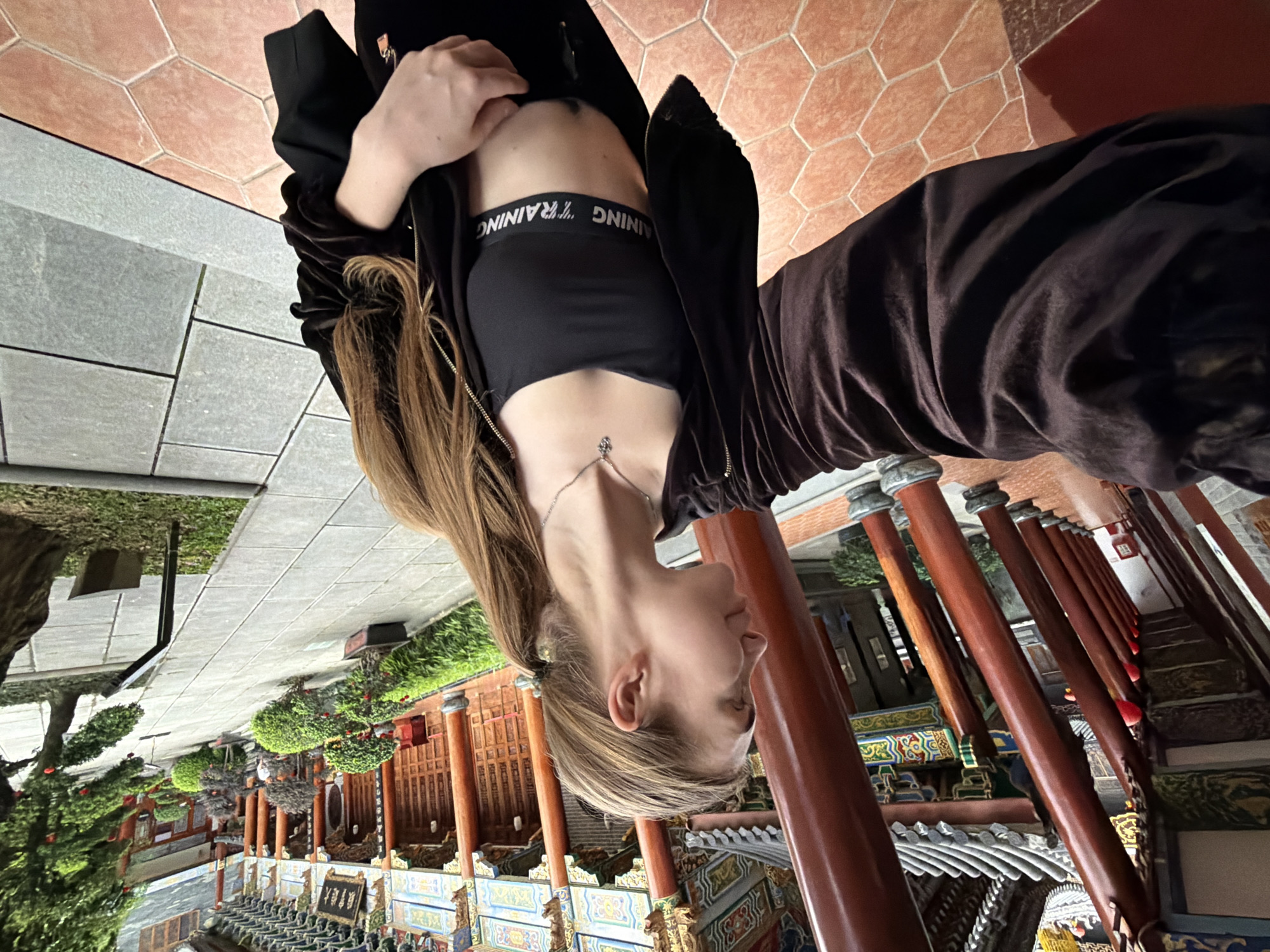 In the city of Longgang District, China, a young woman with long, blonde hair pulled into a ponytail takes a selfie in a courtyard with traditional Chinese architecture. She is wearing a black sports bra with white "TRAINING" lettering and a dark brown velvet zip-up jacket, revealing her midriff. She is also wearing black pants. The background features a covered walkway with large, red wooden pillars and intricate decorative details on the building's roof. Beyond the walkway, a tiled path leads to a garden with manicured bonsai trees and larger trees. The ground in the foreground is tiled with hexagonal terracotta tiles, and further back, a path is laid with large grey square stones. The lighting suggests it is daytime.