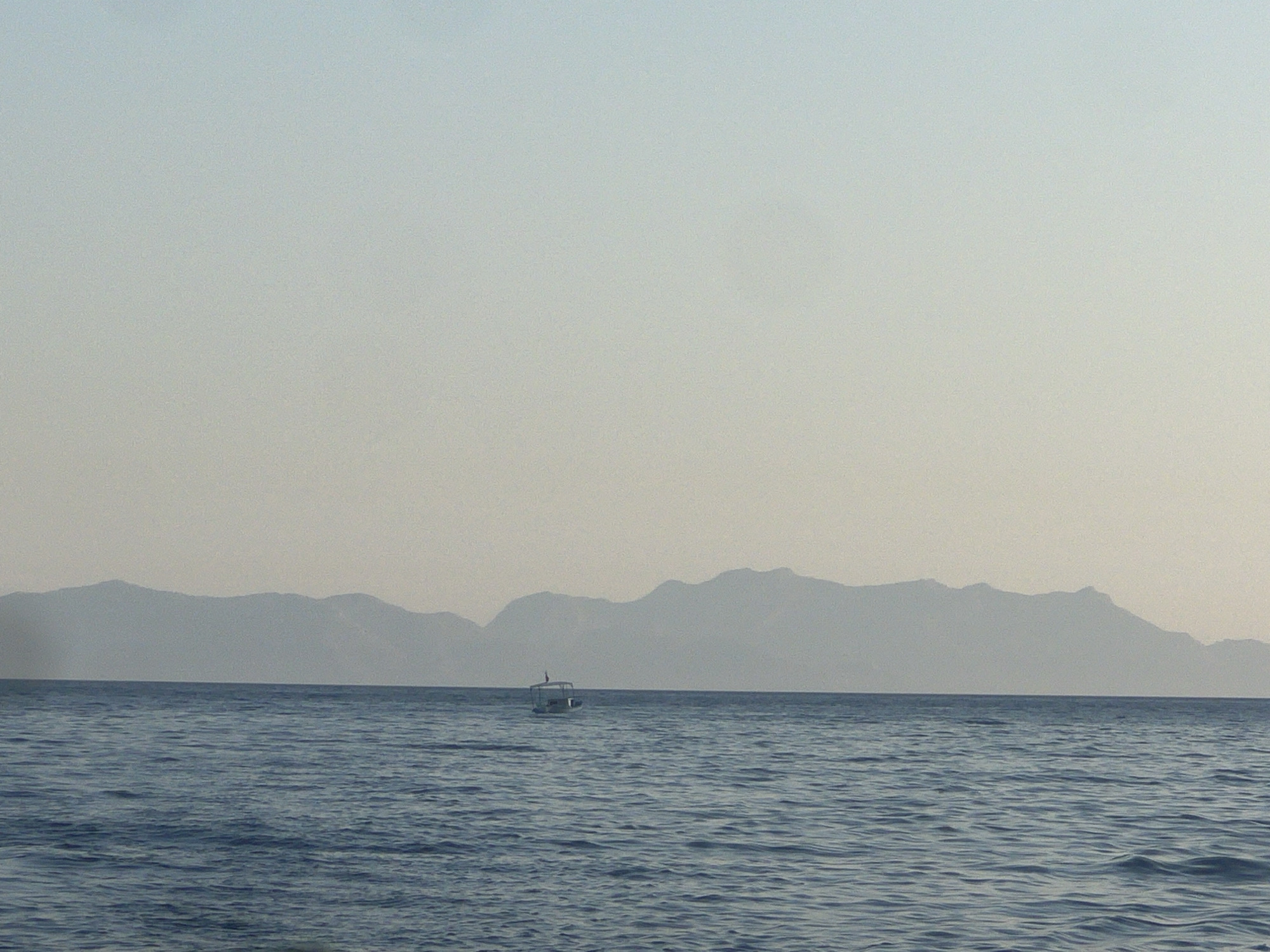 A distant, hazy view of a body of water, likely the sea, stretches across the bottom of the image. The water is a deep blue with gentle ripples. In the middle distance, a small, white boat with a canopy is visible, sailing on the water. The horizon is marked by a range of silhouetted mountains, their peaks softened by a light haze. The sky above is a pale, gradient blue, with a warmer, lighter tone near the horizon. The overall scene suggests a calm, early morning or late afternoon ambiance.