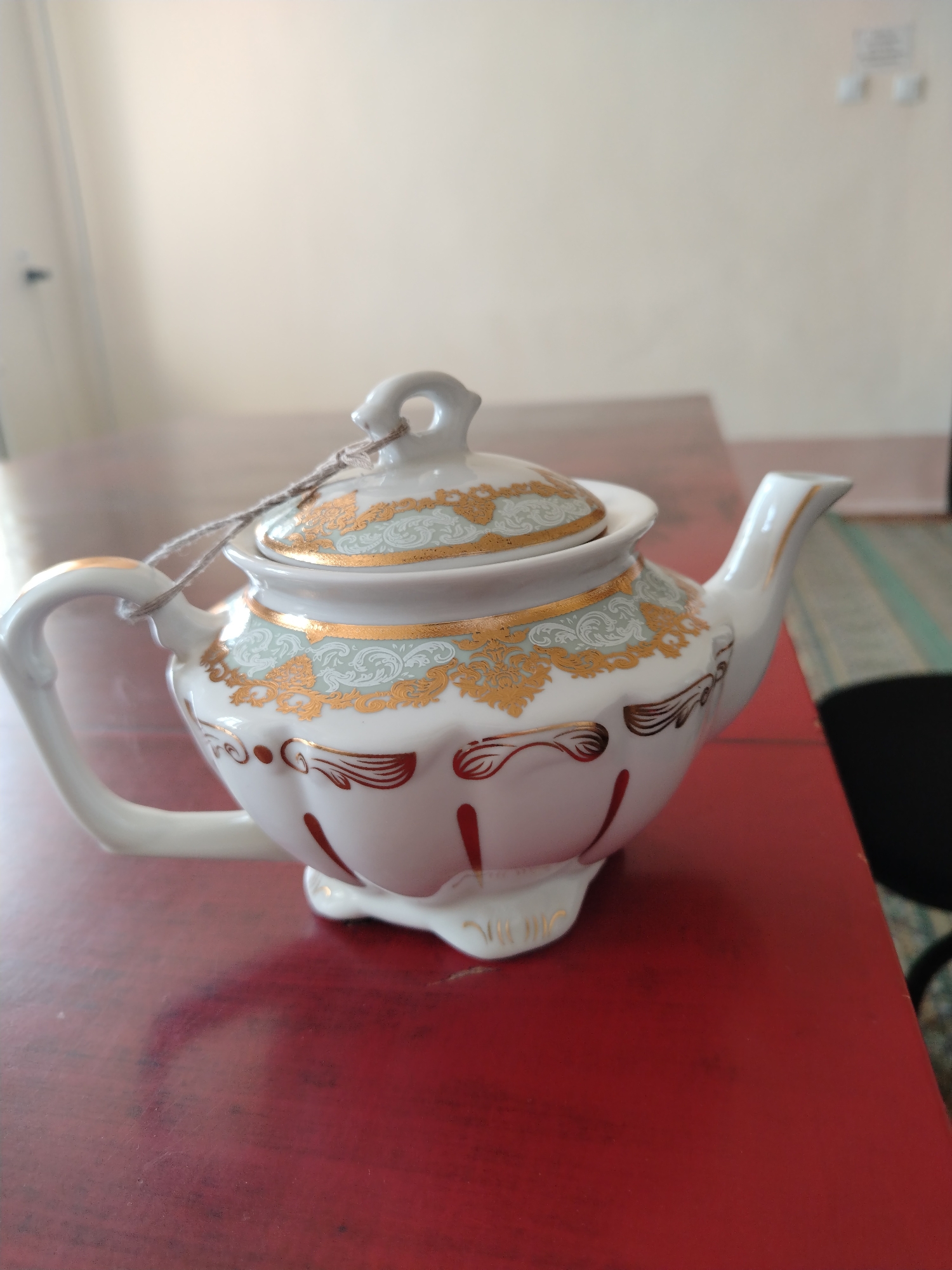 A white ceramic teapot with gold and light green decorative patterns sits on a dark red wooden table. The teapot has a handle and a spout, and a lid secured with a light brown string. The background is a lightly colored wall with a door on the left. The scene is indoors. The image was taken in Nurata, Uzbekistan.