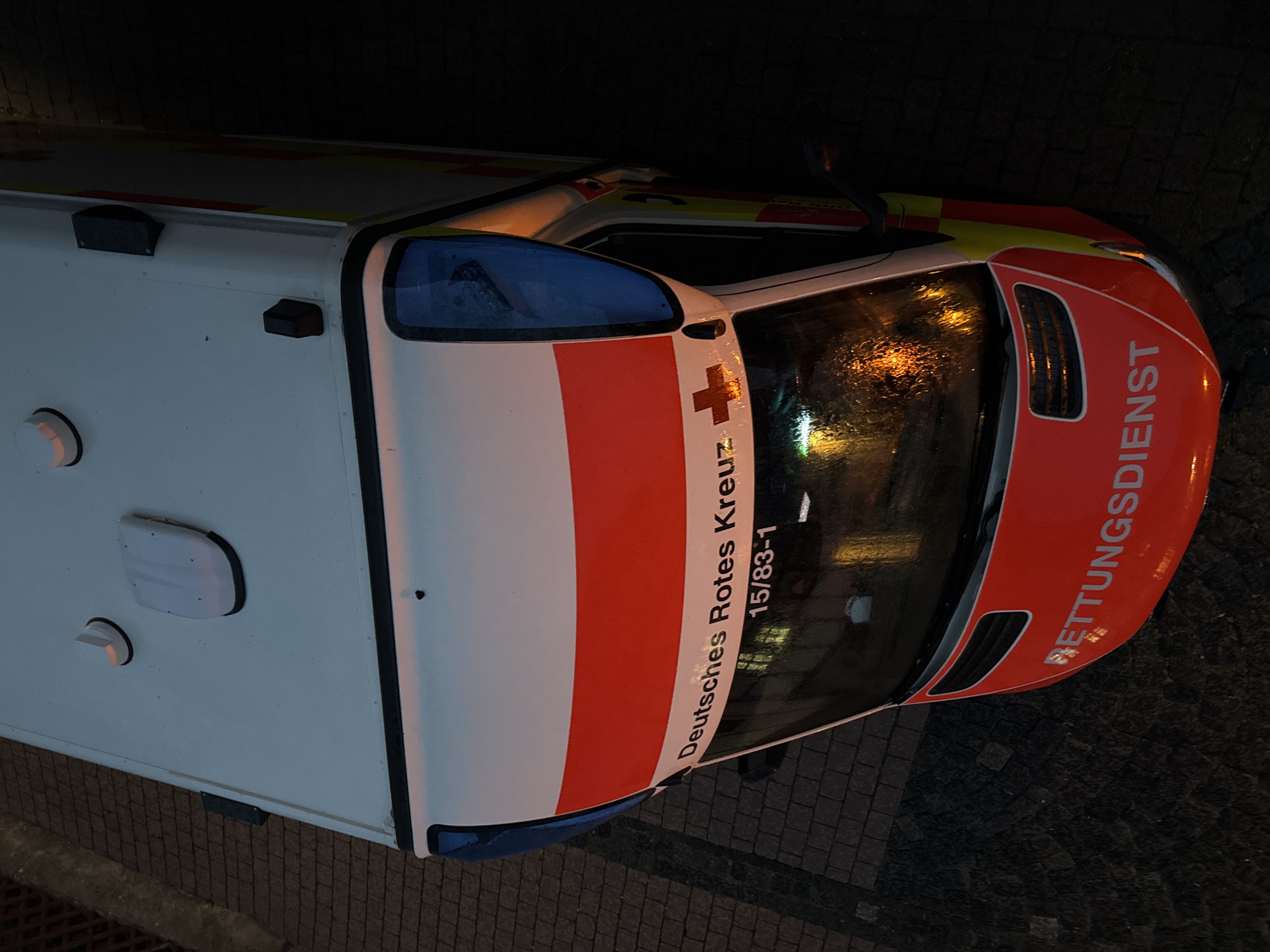 An overhead, eye-level, close-up shot captures the front portion of a German Red Cross ambulance parked on a cobblestone street at dusk or dawn. The white roof of the ambulance is visible, with two dome-shaped lights and a rectangular black component. A prominent red and white stripe runs horizontally across the vehicle's front, with a red cross emblem on the left side. Text below the stripe reads "Deutsches Rotes Kreuz 15/83-1". The windshield is wet and reflects the orange glow of streetlights, indicating recent rain. The lower front of the ambulance is bright red with a white stripe, and the word "RETTUNGSDIENST" is visible in white lettering. The surrounding environment is dark, with hints of a brick wall on the right and a tiled roof on the left.