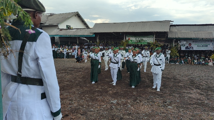 The image shows a group of people in white and green uniforms standing in formation in an open area in front of a building in the city of Keshero, Democratic Republic of the Congo. The building has a corrugated metal roof and appears to be partially constructed. There are other people watching from the sidelines. One person in the foreground is wearing a green beret and white and green uniform, standing facing to the left of the frame. The people in the formation are performing a drill or marching routine. The ground is dirt and there is foliage in the foreground. There is a banner hanging on the building that says "G.T.K." and "KOKO KIVU". There is also a poster with a picture of a man on it. The photo appears to have been taken during a formal event, perhaps a ceremony or parade. The mood is one of order and formality. The event appears to be taking place in a public space, perhaps a schoolyard or a community gathering area. The event appears to be related to the "G.T.K.", which could be a school, organization, or community group.