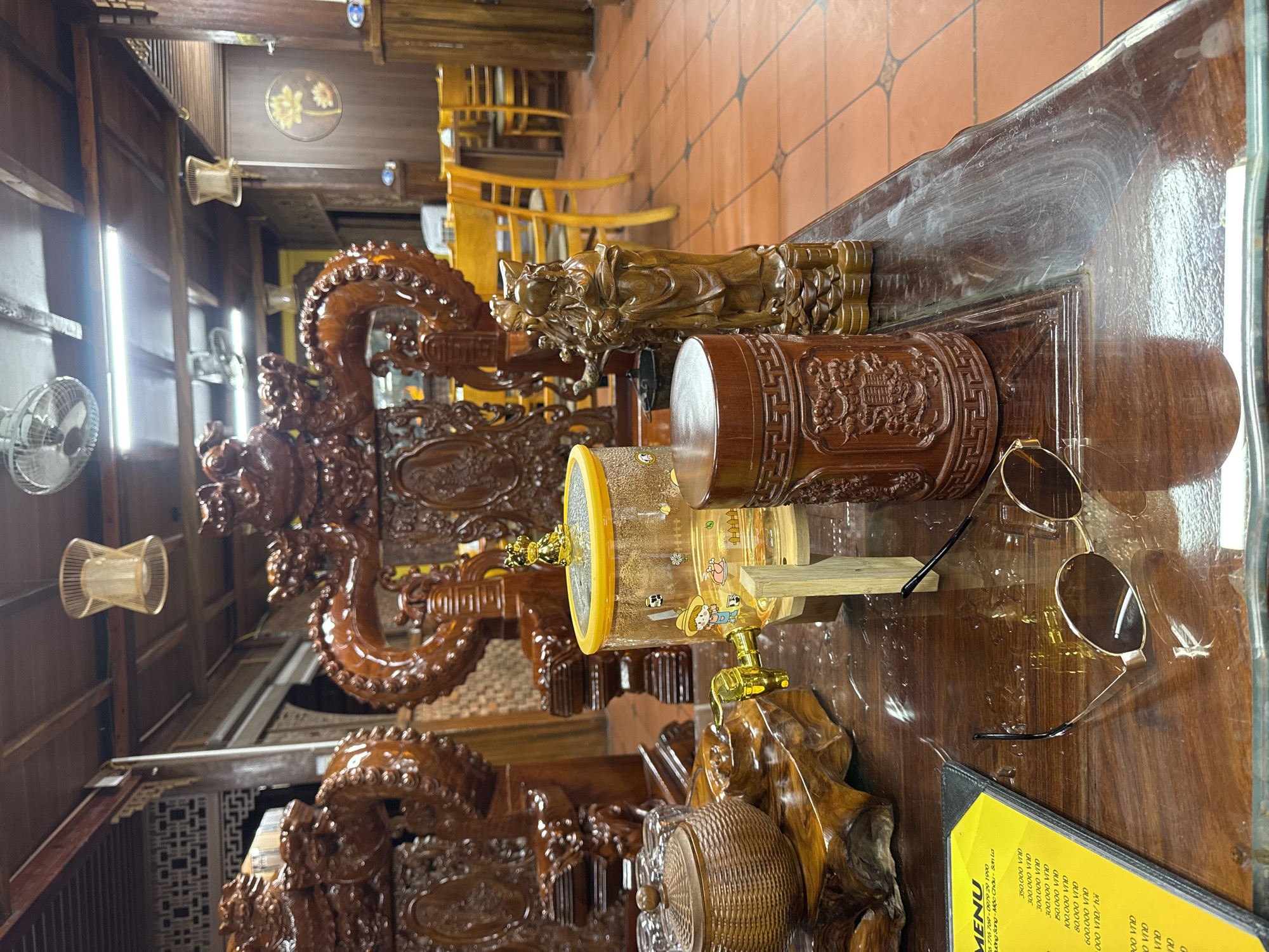 The image depicts a wooden table in what appears to be a restaurant or dining area in Mộc Châu, Vietnam. The table is set with various decorative and functional items. Prominently featured are ornate wooden carvings, possibly depicting dragons, and two wooden figurines of individuals. On the table, there's a clear beverage dispenser with a gold spigot, decorated with cartoon-like images. To the left of the dispenser, a woven basket with a lid and a set of small glasses are visible. Also on the table are a pair of sunglasses and a yellow menu. The background reveals more wooden furniture, including chairs and tables, and traditional architectural elements such as lattice work and ceiling fans. The floor is tiled in a terracotta color. The overall scene suggests a traditional Vietnamese setting with an emphasis on wood craftsmanship and possibly a focus on beverages.
