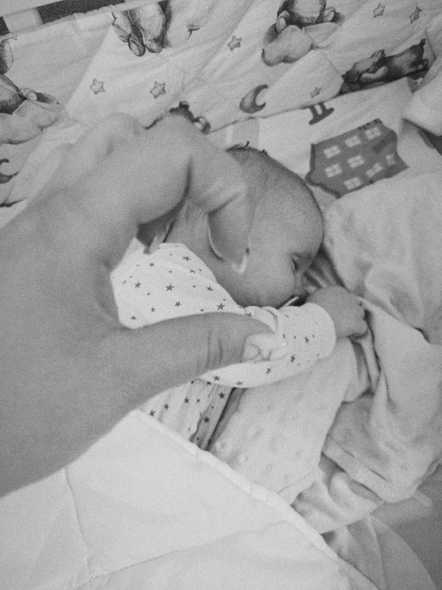 A black-and-white, high-angle, medium shot photograph shows a baby sleeping on patterned bedding with an adult hand hovering above. The baby is lying on its side, wearing a star-patterned pajama. The hand is positioned on the left, partially obscuring the baby, appearing as if someone is placing their hand over the baby. The bedding has a cartoonish pattern of toys and geometric shapes. The location and presence in Tobolsk, Russia cannot be determined from the visual information.