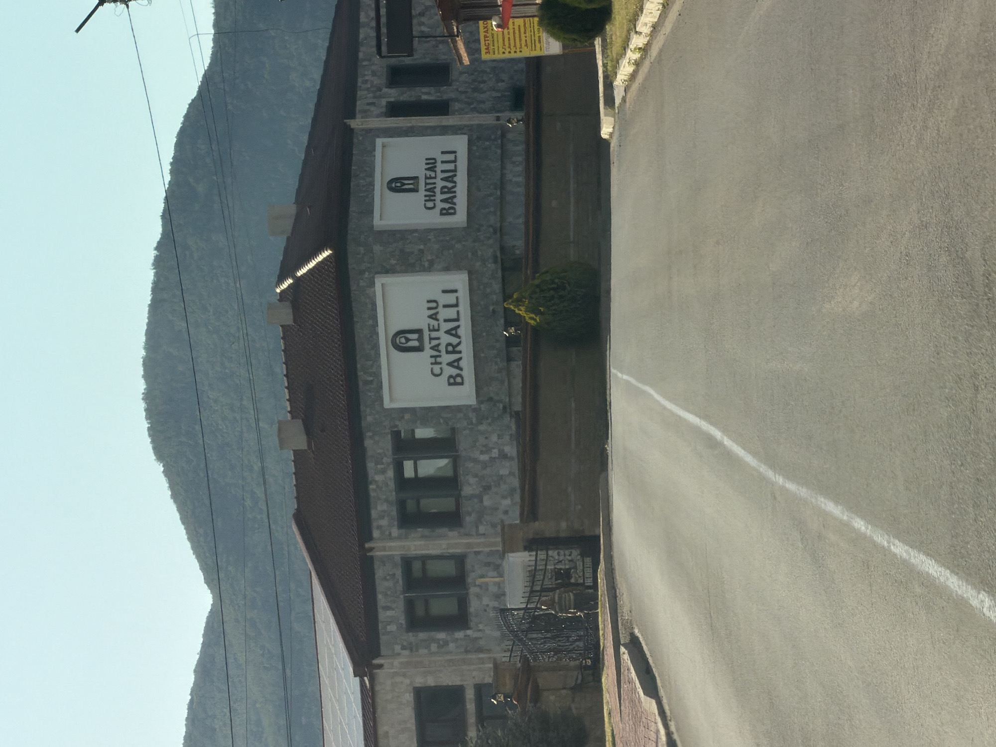 This image depicts the entrance to Chateau Baralli in Kamchiya, Bulgaria. The chateau is a two-story building with a dark tiled roof and a facade of light-colored stone tiles. Two large signs on the facade read "CHATEAU BARALLI" with an emblem of a wine glass above a person. The building is set against a backdrop of green, tree-covered mountains under a clear blue sky.
In front of the chateau is a black wrought-iron gate. A person, seen from behind and wearing a striped shirt and dark pants, is standing near the gate. A paved road leads directly towards the chateau entrance. The road has a white line marking the center. To the right of the road, there is a yellow sign with Bulgarian text, partially obscured by bushes. The overall scene suggests a business establishment, likely a winery or hotel, situated in a scenic, possibly rural, location.