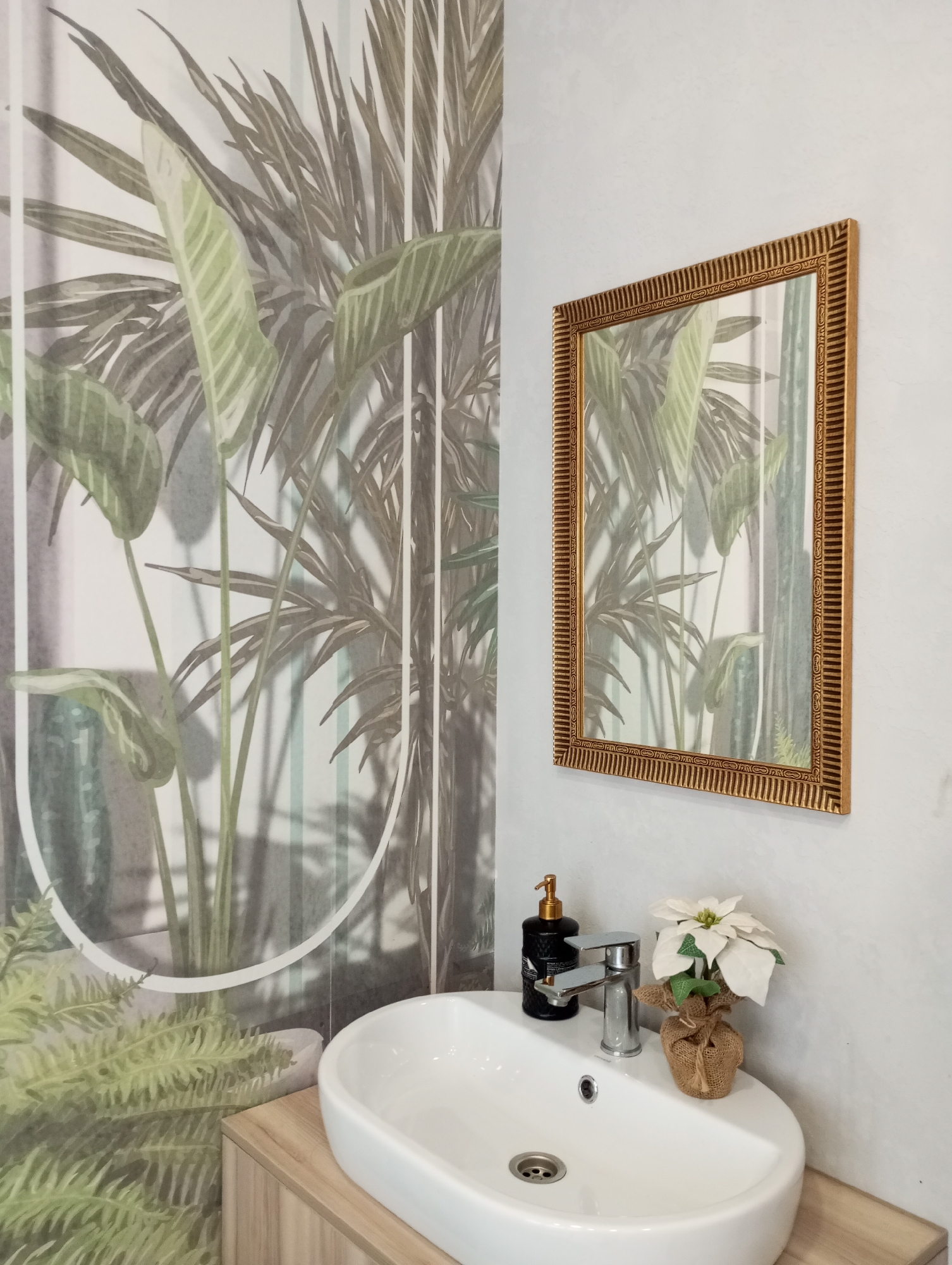 A bathroom interior is shown. A white, oval-shaped sink is set into a light wood-colored cabinet. A silver faucet and a black soap dispenser are located on the sink surface, along with a small artificial white poinsettia plant in a burlap-wrapped pot. A gold-framed square mirror hangs on the wall above the sink. The wall on the left is decorated with a wallpaper featuring an image of green tropical plants. The walls appear to be a light gray or off-white color with a textured finish. The location is Izhevsk, Russia.