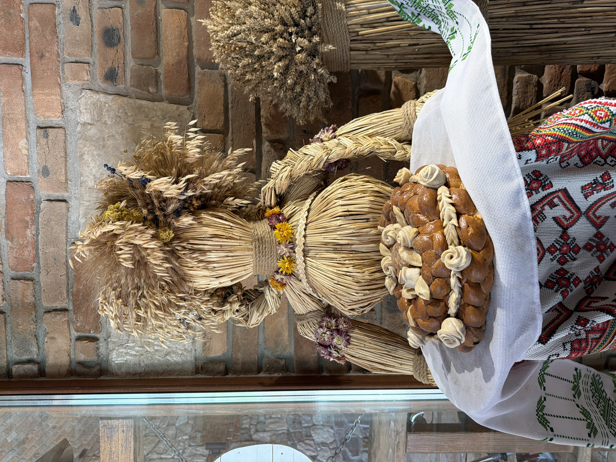 An indoor scene depicts a straw effigy dressed in traditional Ukrainian attire, seated before a brick wall. The effigy is adorned with dried flowers and grains, and its hands are positioned on a decorative braided bread, which rests on a white tablecloth with embroidered borders. To the right, another straw decoration with wheat stalks stands upright. The setting appears to be within a building, possibly a cultural center or a home, in the city of Velykyi Khodachkiv, Ukraine.