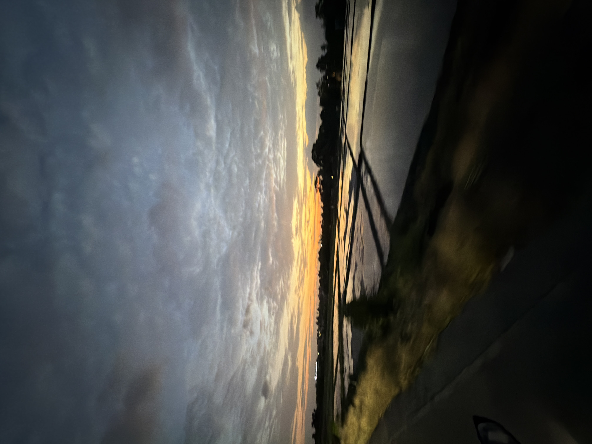 The image captures a sunset viewed from a moving vehicle near North Kuta, Indonesia. The left side of the frame features a dramatic sky with layered, gray clouds. An orange and yellow glow from the setting sun extends horizontally across the middle of the frame, highlighting the horizon line and the tops of trees. A road runs along the right side, with roadside vegetation visible. Parts of the vehicle's interior are visible in the foreground at the bottom. The lighting conditions suggest dusk.
