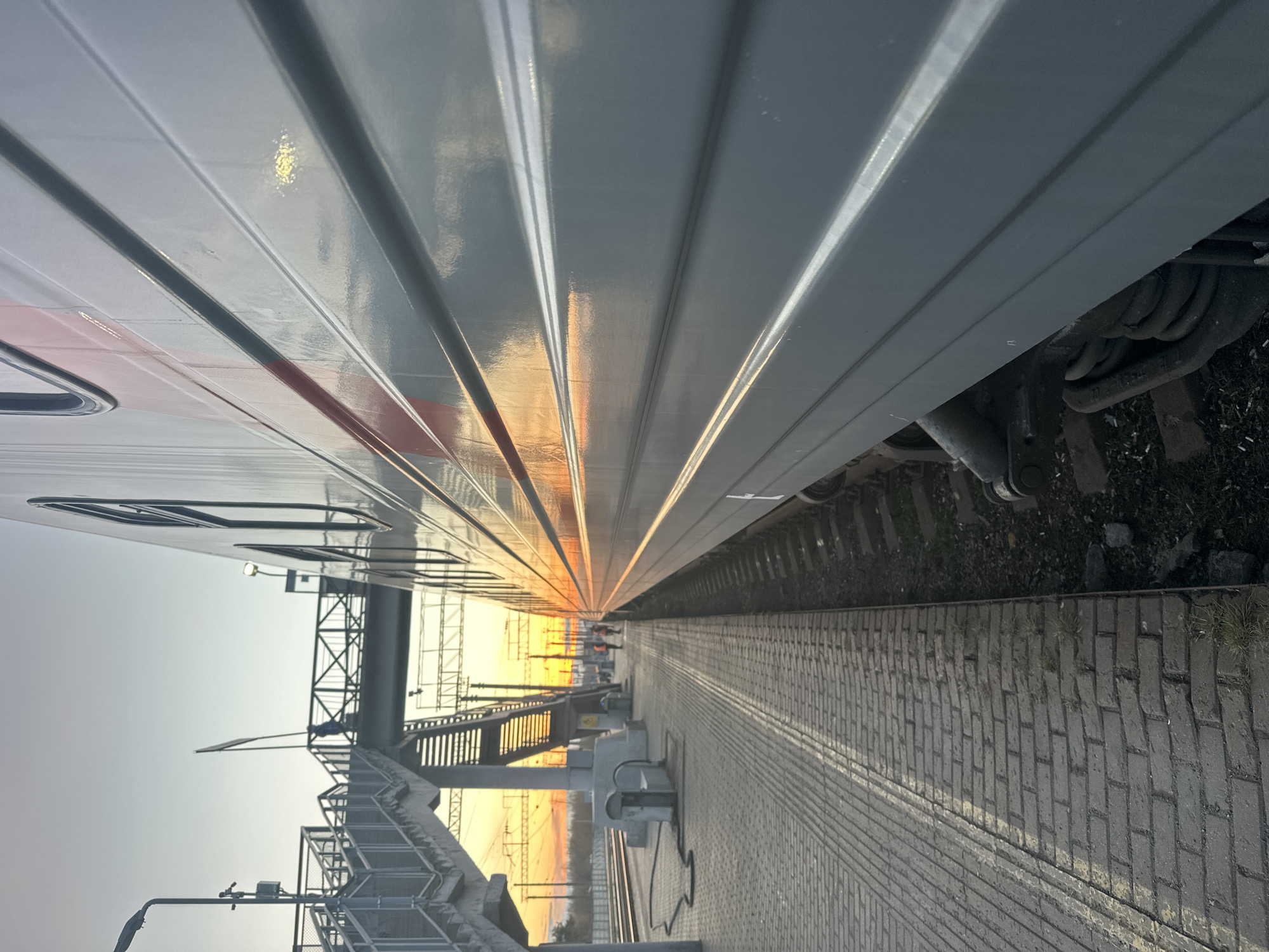 The image depicts a view along a train platform at sunrise or sunset, likely in Agryz, Russia. A passenger train is stationed to the left. The train's exterior is light gray with vertical ridges and some red accent. A raised pedestrian walkway bridge spans the tracks in the distance. The platform is paved with rectangular bricks. The lighting suggests either early morning or late evening. Train tracks are visible between the platform and the train. Utility poles and wires are present in the background. There are silhouettes of people standing on the platform in the distance.