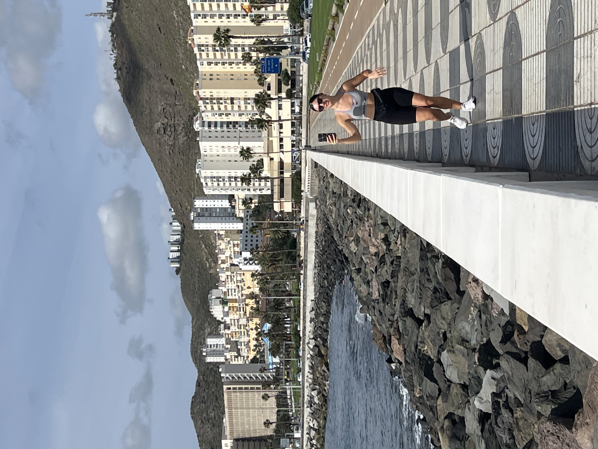 A full-length view shows a woman taking a selfie on a pedestrian walkway in Las Palmas, Spain. The woman, wearing a grey athletic top, black shorts, and white sneakers, stands facing the camera while holding her mobile phone in her right hand. She has her left arm outstretched. She is also wearing sunglasses.
The pedestrian walkway has a pattern of alternating grey and white semi-circles. A white concrete wall separates the walkway from a rocky beach and the ocean. Across the walkway, a boulevard is lined with palm trees and buildings that appear to be apartments and businesses. A large mountain looms behind the buildings. The sky is partly cloudy.
The location is Las Palmas, Spain.
