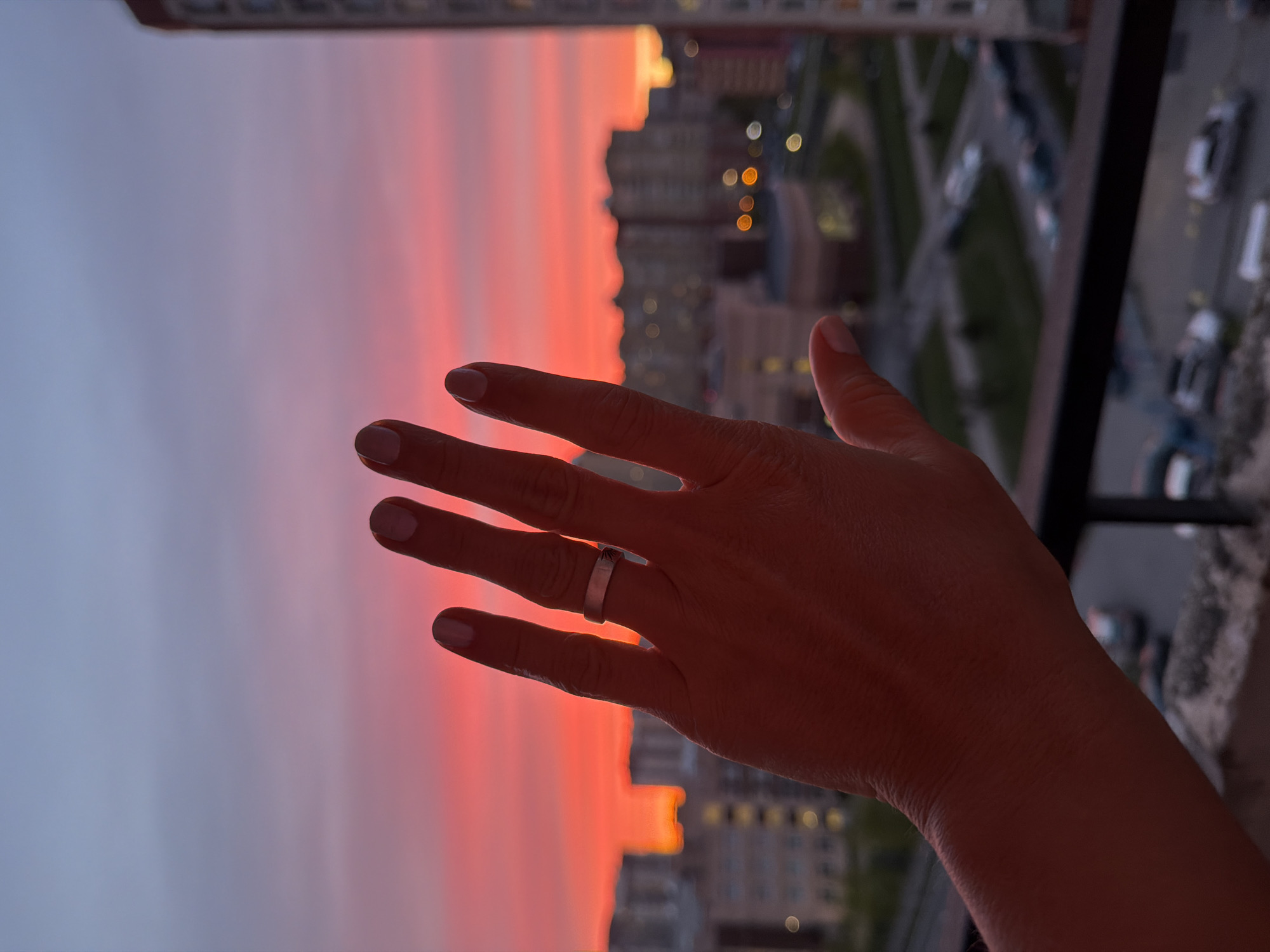 A vertical photograph shows a woman's right hand, wearing a silver ring on her ring finger, extended against a twilight sky. The sky transitions from grey near the top of the frame to vibrant shades of red and orange closer to the horizon. Beyond the hand and the sky, there is a blurry cityscape featuring buildings with illuminated windows, suggesting that it is dusk or night. The scene includes streets with parked cars and patches of green, indicating urban landscaping. Based on the architecture and urban layout, the location may be Saint Petersburg, Russia.