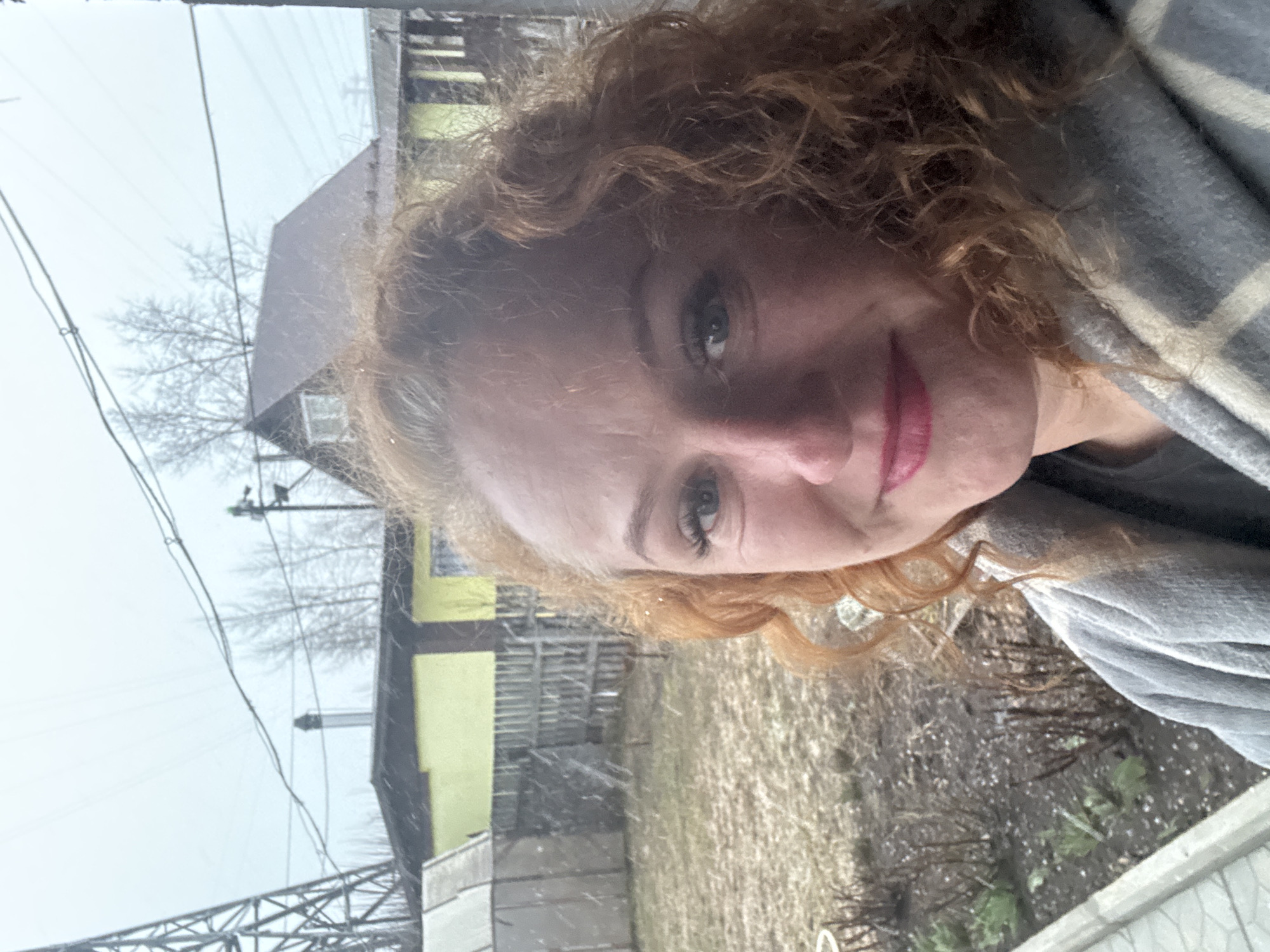 A selfie of a woman with curly red hair, light skin, and red lipstick, wearing a gray and white patterned blanket. She is facing the camera with a slight smile. The background shows a cloudy day with a building painted yellow and black, partially obscured by bare trees and power lines. Ground vegetation is visible. Based on the presence of buildings and utilities, it is likely a residential area. Given the prompt's context, this is plausibly Chaikovsky, Russia.