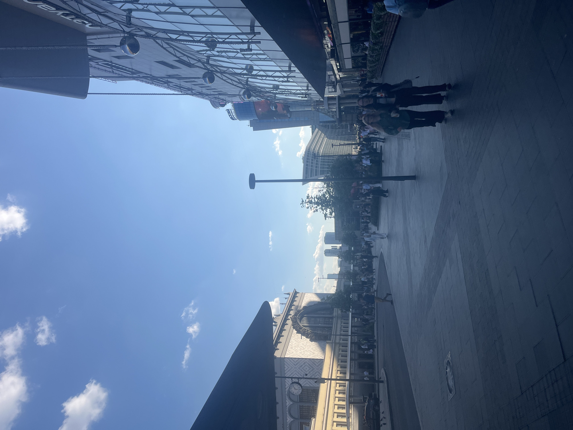This image captures an outdoor urban scene in Moscow, Russia, specifically the area surrounding Belorussky Railway Station. The sky is bright blue with scattered white cumulus clouds.
On the left, a large, light-colored architectural structure, identifiable as Belorussky Railway Station, dominates the mid-ground. It features a grand central arch, ornate patterned stonework on its upper facade, and a round clock on a lower section to the left of the main entrance. Below the station facade, a paved area with a dark vehicle partially visible and several pigeons on the ground suggests a drop-off or taxi zone.
On the right, a modern, multi-story building with a reflective glass facade extends vertically. This building features a dark, curved canopy overhanging the pedestrian area. Numerous large, dark spherical ornaments are suspended by chains along the building's exterior. Cyrillic text, including "ЭТУАЛ" and a partial "СКИЙ," is visible on the upper sections of this building. The ground level includes establishments with outdoor seating and planter boxes.
The central and foreground areas consist of a wide, gray pedestrian plaza paved with rectangular tiles. Numerous individuals are walking across this plaza in various directions. In the immediate foreground, two women walk side-by-side towards the viewer, both holding cups. Further back, a man in light-colored trousers and a dark shirt walks away from the viewer. Another person in a long white garment walks further to the right. A tall, black street light pole stands in the mid-ground to the left of the pedestrian flow.
In the background, several modern high-rise buildings are visible, including one with a distinctive curved or angled design, contributing to the city's skyline. Scattered trees and foliage provide some greenery. The scene is brightly lit by sunlight, casting shadows indicative of a clear daytime environment.