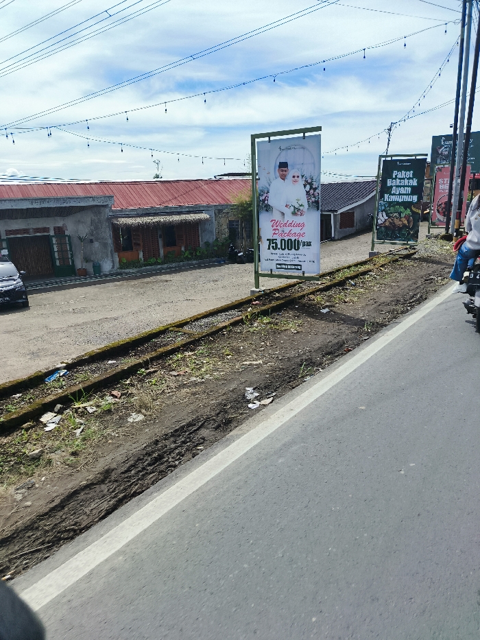The image shows a paved road with a white line separating the lanes. There is a dirt shoulder on the side of the road with overgrown grass and weeds. In the background there are buildings with a red tiled roof. On the side of the road, a man is riding a motorbike and a large billboard with a wedding package advertisement is standing. To the right of the billboard is another sign advertising a "Paket Bakakak Ayam Kampung." The location is in Rancabali, Indonesia. The coordinates are -7.122 latitude and 107.427 longitude.