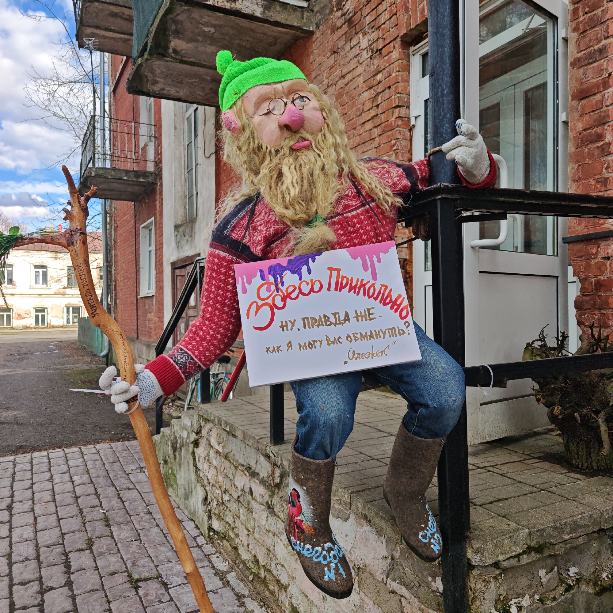 The image depicts a humorous, makeshift character placed on a brick ledge or step, potentially in the city of Rostov, Russia, given the Cyrillic script on the sign and the stick. The character, likely a scarecrow or decorative figure, is dressed in clothing that includes a red and black patterned sweater, blue jeans, and what appear to be felt boots with writing and designs. It wears a green hat and glasses, and features a face with a pronounced nose and beard. The figure holds a wooden stick in one hand and a sign in the other. The sign is brightly decorated and contains a message in Russian, "Здесь Прикольно" (roughly translated as "It's fun here"), followed by "Ну, правда же. Как я могу вас обмануть? «Олежек»" (which can be understood as "Well, really. How can I deceive you? «Olezhek»").
The figure is positioned near a building with red brick walls, a black metal railing, and a white door. The building is part of an urban setting, as seen by the pavement beneath and other buildings in the background. The overall impression is a whimsical attempt to attract attention or create a welcoming atmosphere.