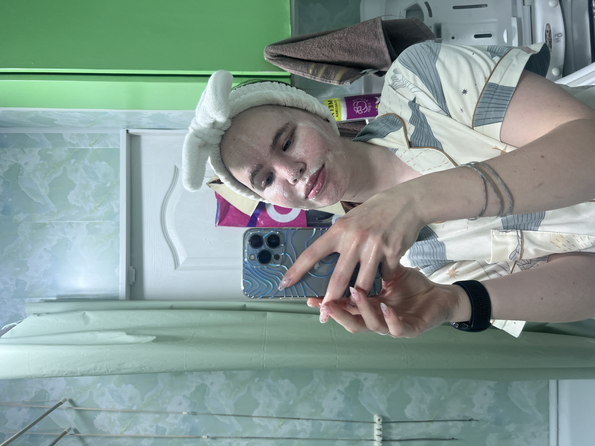 A person, likely female, is captured in a mirror selfie while performing a skincare routine in an indoor setting, possibly in Chaikovsky, Russia. They have a white face mask or cleanser applied to their skin and are wearing a white fluffy headband with bunny ears. Their pajamas feature a pattern of grey and blue geometric shapes and small stars.
The individual is holding a smartphone, which appears to be an iPhone with multiple camera lenses, encased in a clear cover with blue wavy lines. A black smartwatch adorns their left wrist, and silver-colored bracelets are visible on their right arm.
The background consists of a bright green wall or shelf at the top, transitioning to a lighter wall with a subtle marbled pattern. A white door or cabinet is also present. A brown towel hangs in the upper right. Partially visible purple and pink product packaging behind the person clearly displays the Cyrillic word "МАСКА" (MASK) and a brand name, possibly "MEEYA". Metallic bars, possibly from a drying rack, are seen towards the bottom of the frame. The image is oriented 90 degrees clockwise.