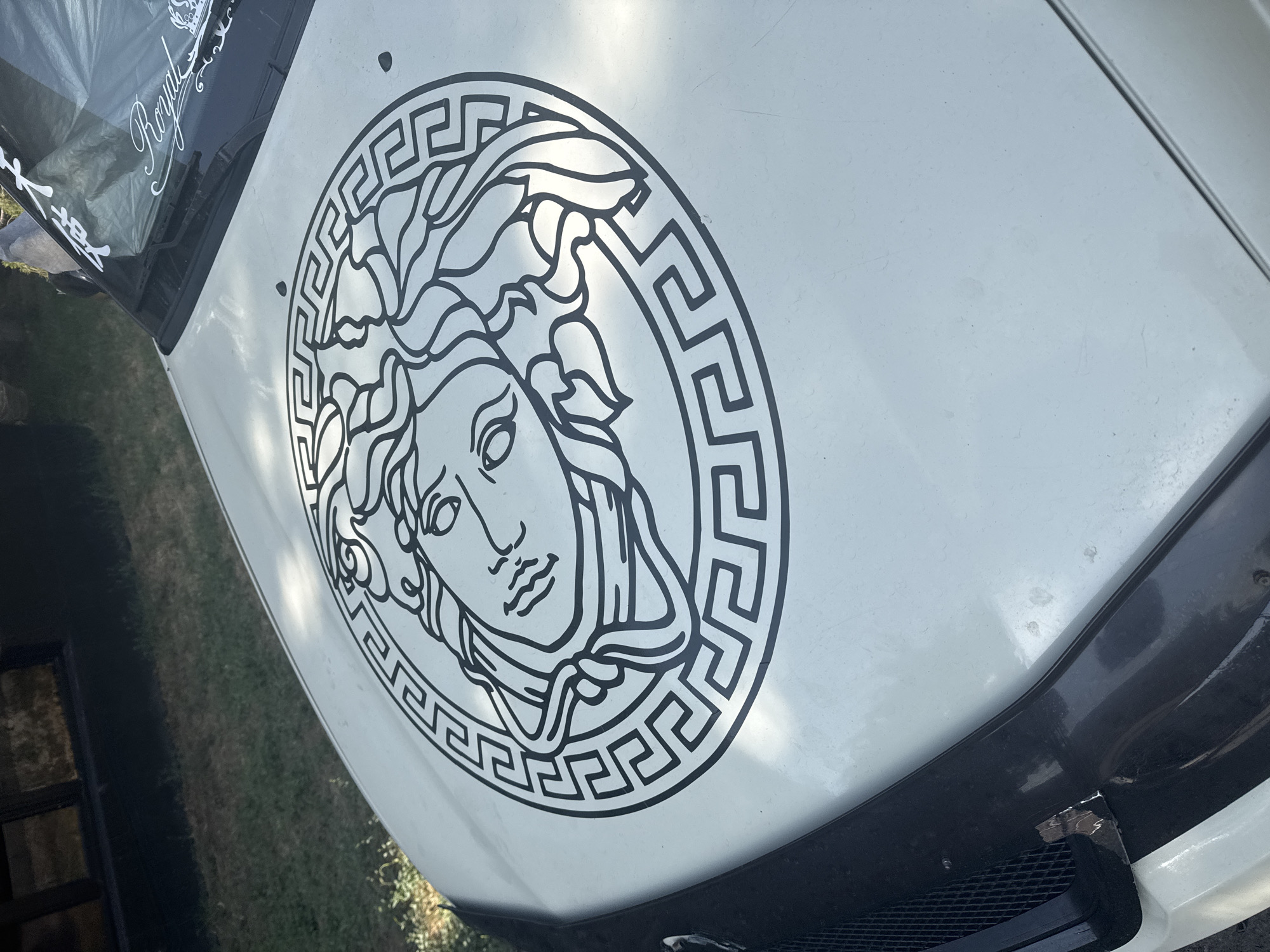 The image shows a close-up, high-angle view of the hood of a light-colored car. A large, black decal of Medusa's head within a Greek key border covers the center of the hood. The car is parked outdoors, with green grass and a dark-colored building or structure visible in the background. Sunlight casts shadows and highlights on the car's surface. In the upper right corner of the windshield, white decals are visible. The overall condition of the car's paint appears somewhat aged, with minor marks and scuffs. No people are visible in the image. The location is not specified in Bishkek City, Kyrgyzstan.