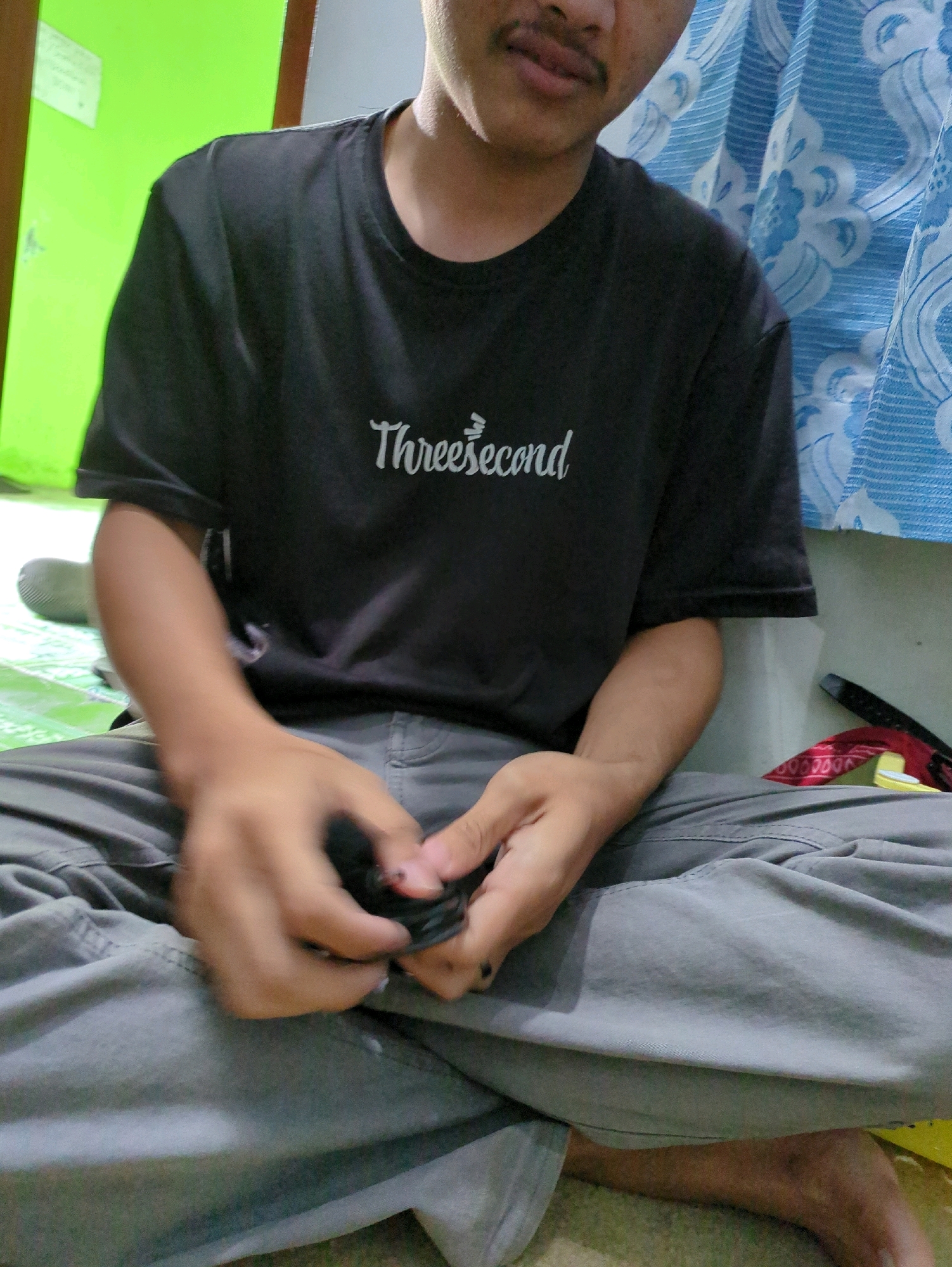 A person is sitting cross-legged on the floor in what appears to be a room in Gumpang, Indonesia. The person is wearing a black t-shirt with "Threesecond" written in white, stylized text, and grey pants. Their hands are engaged in an action, holding a small, dark object. The background includes a bright green wall on the left and a blue patterned curtain on the right. A shoe can be seen to the left of the person. The lighting suggests an indoor environment.