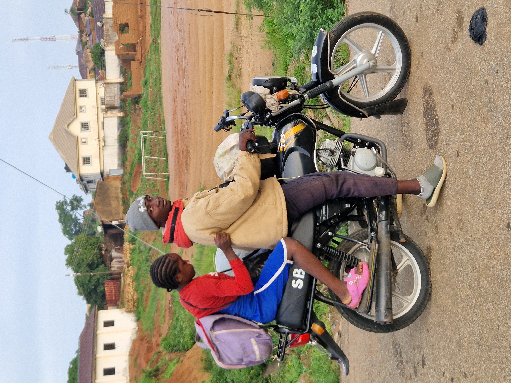 In Kabong, Nigeria, a young boy wearing a gray beanie, a tan jacket with a red collar, and dark pants rides a black motorcycle. Seated behind him is a young girl with braided hair, wearing a red long-sleeved shirt, a blue dress, and pink sandals. She has a purple backpack slung over her shoulder. The motorcycle has yellow and black markings and the letters "SB" are visible on the seat. They are on an asphalt road, with a grassy area and dirt paths on either side. In the background, there are several buildings, including a light-colored two-story house, and some unfinished brick structures. A white soccer goal is visible in the middle ground. The sky is overcast.