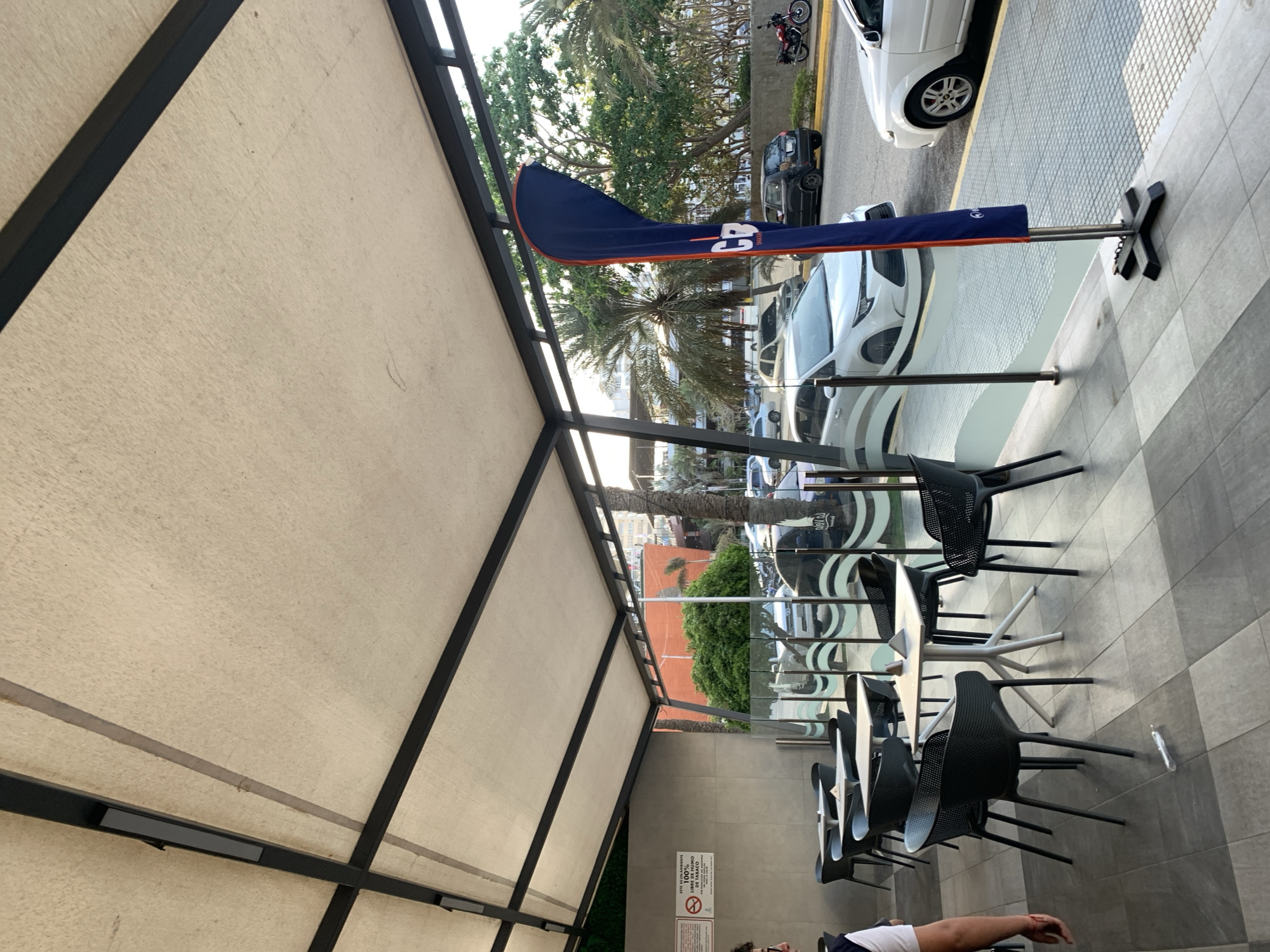 The image shows an outdoor seating area with a view of a street in Porlamar, Venezuela. The seating area is covered by a beige awning with a black frame. There are several black chairs and white tables arranged in rows. A blue flag with white letters, possibly a brand logo, stands on a pole near the edge of the seating area. The street view includes cars, motorcycles, palm trees, and other vegetation. The flooring of the seating area is tiled in gray. A person's arm is visible in the lower right corner of the image. A sign with text in Spanish is mounted on a wall near the back of the seating area.