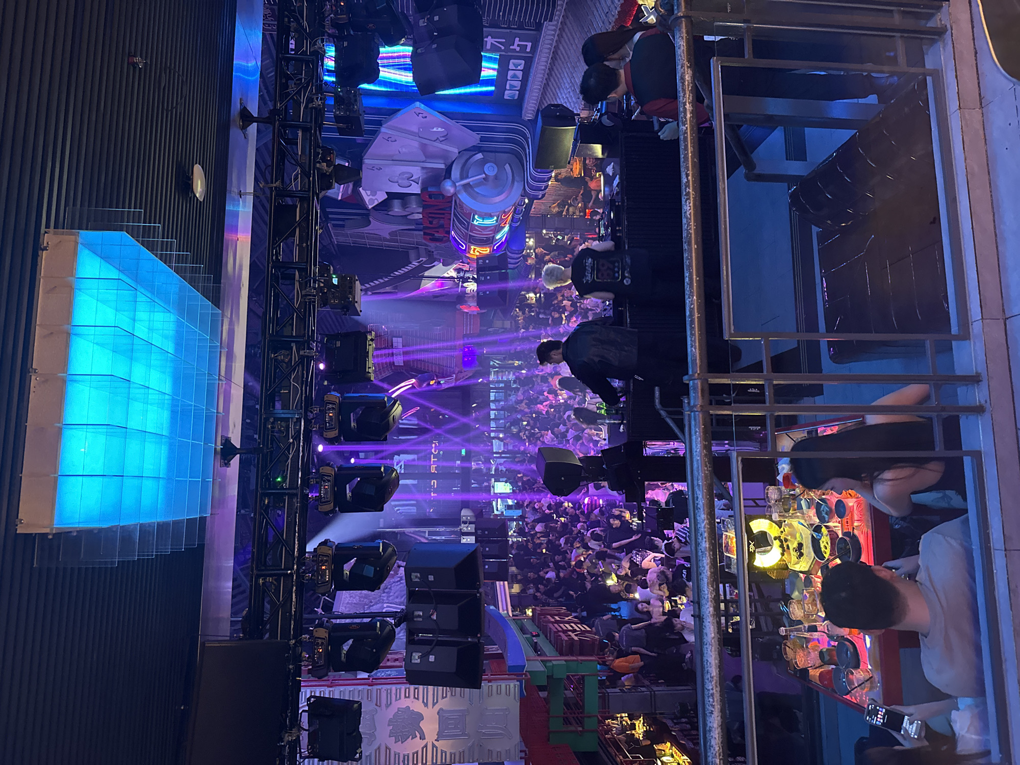 The image depicts the interior of a crowded nightclub in Thủ Đức, Vietnam. The scene is illuminated by vibrant purple and blue lights emanating from stage lighting and decorative elements. A DJ booth is visible in the foreground, with two DJs appearing to be performing. A large crowd of people is gathered on the dance floor, many of whom are dancing or socializing. Several tables are set up in a section of the club where patrons appear to be seated and drinking. The overall atmosphere suggests a lively and energetic nighttime entertainment venue. The architecture includes a multi-level design with a railing in the foreground, suggesting a vantage point overlooking the main area. Various speakers and lighting equipment are suspended from the ceiling. Decorative elements, including what appear to be playing cards and a slot machine facade, contribute to a casino-themed aesthetic.