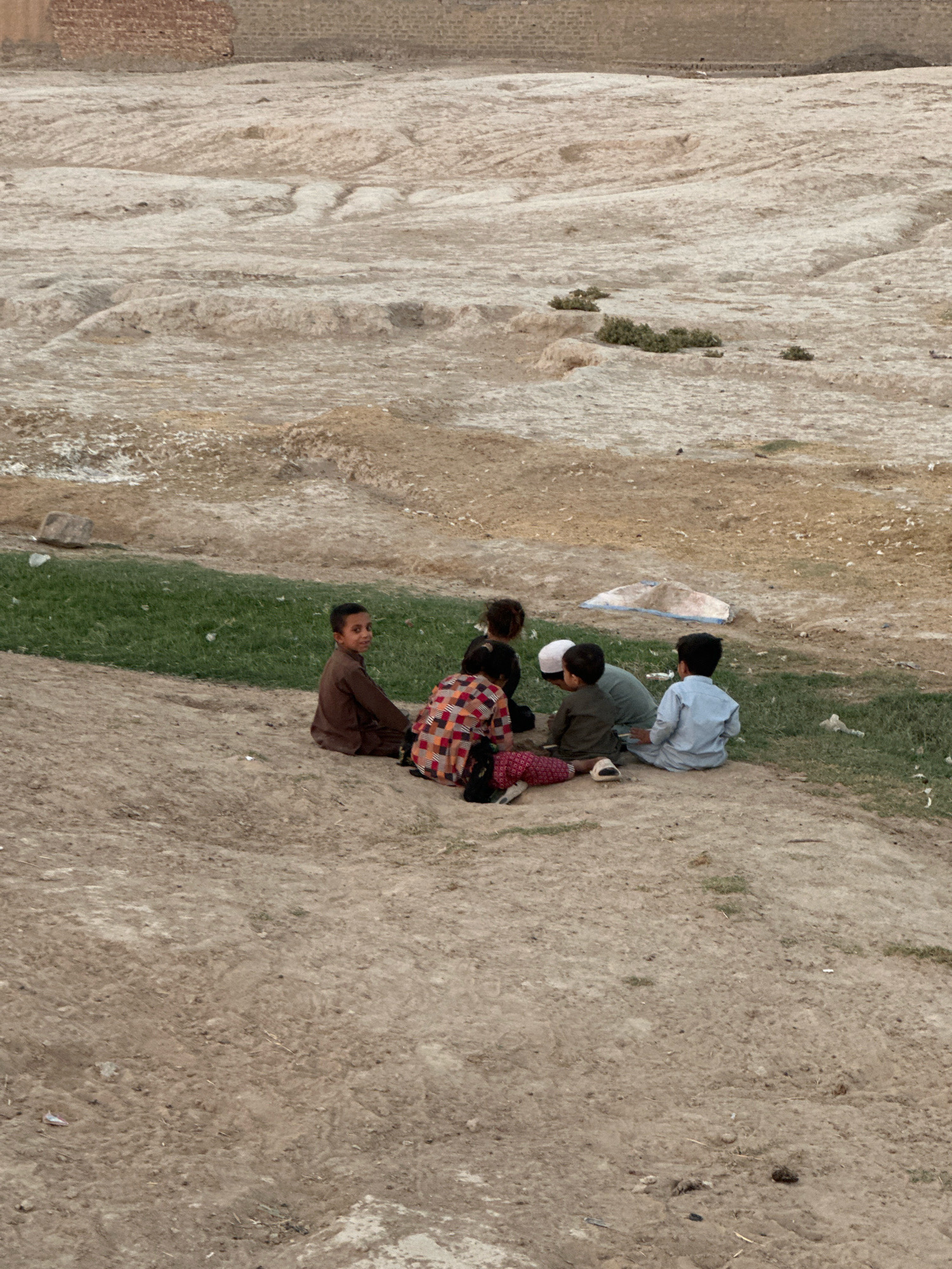 A group of five children are gathered in a circle on a dirt path in Kuchlak, Pakistan. The children appear to be playing or conversing. The ground is a mix of dirt and sparse grass, with a rough, textured terrain in the background. The children are dressed in casual clothing, and the overall atmosphere seems peaceful.