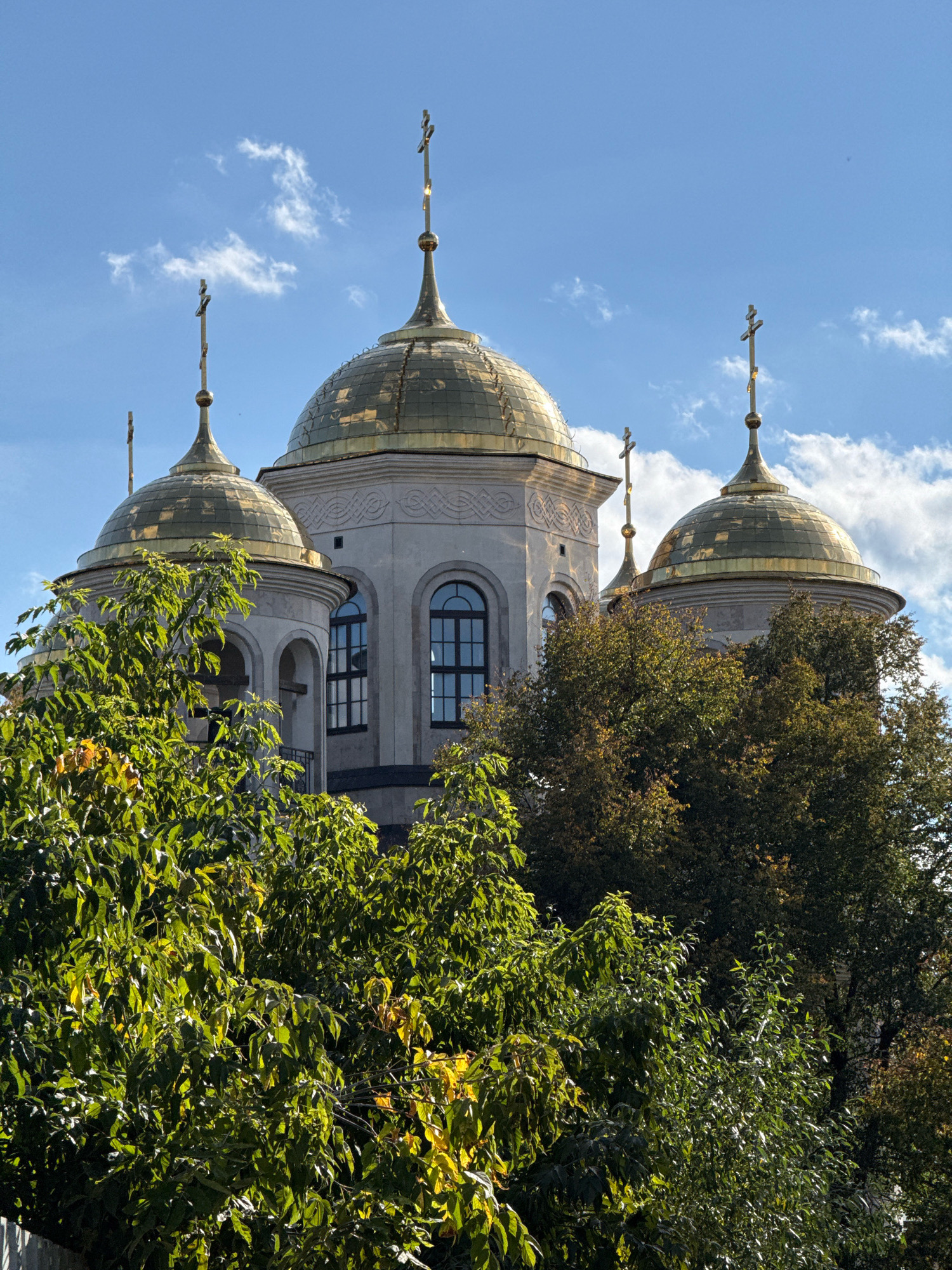 An outdoor, eye-level, medium shot captures the golden domes and crosses of a church, partially obscured by the lush green foliage of trees in the foreground. The sky is a vibrant blue, dotted with fluffy white clouds, suggesting a bright, sunny day. The church's architecture features light-colored stone walls and arched windows. The image appears to be taken in the city of Zvenigorod, Russia, where numerous historical churches with similar golden domes can be found. No people are visible in the scene.