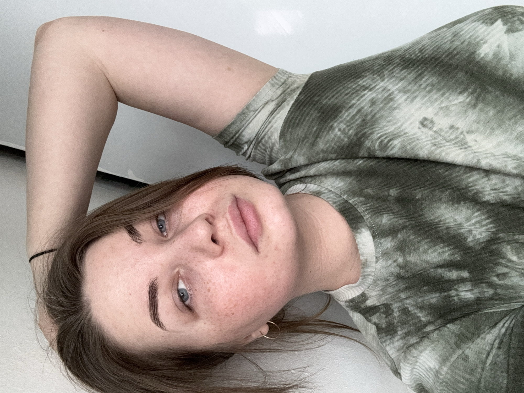 A light-skinned woman with freckles is lying on a light surface, possibly a bed or floor, in an indoor environment. She is wearing a green and white tie-dye t-shirt and a small gold hoop earring is visible on her left ear. Her hair is brown, with a black hair tie visible. The background is plain white. The location of the image is not determinable without additional information. The city of Chaikovsky, Russia cannot be determined from this image.