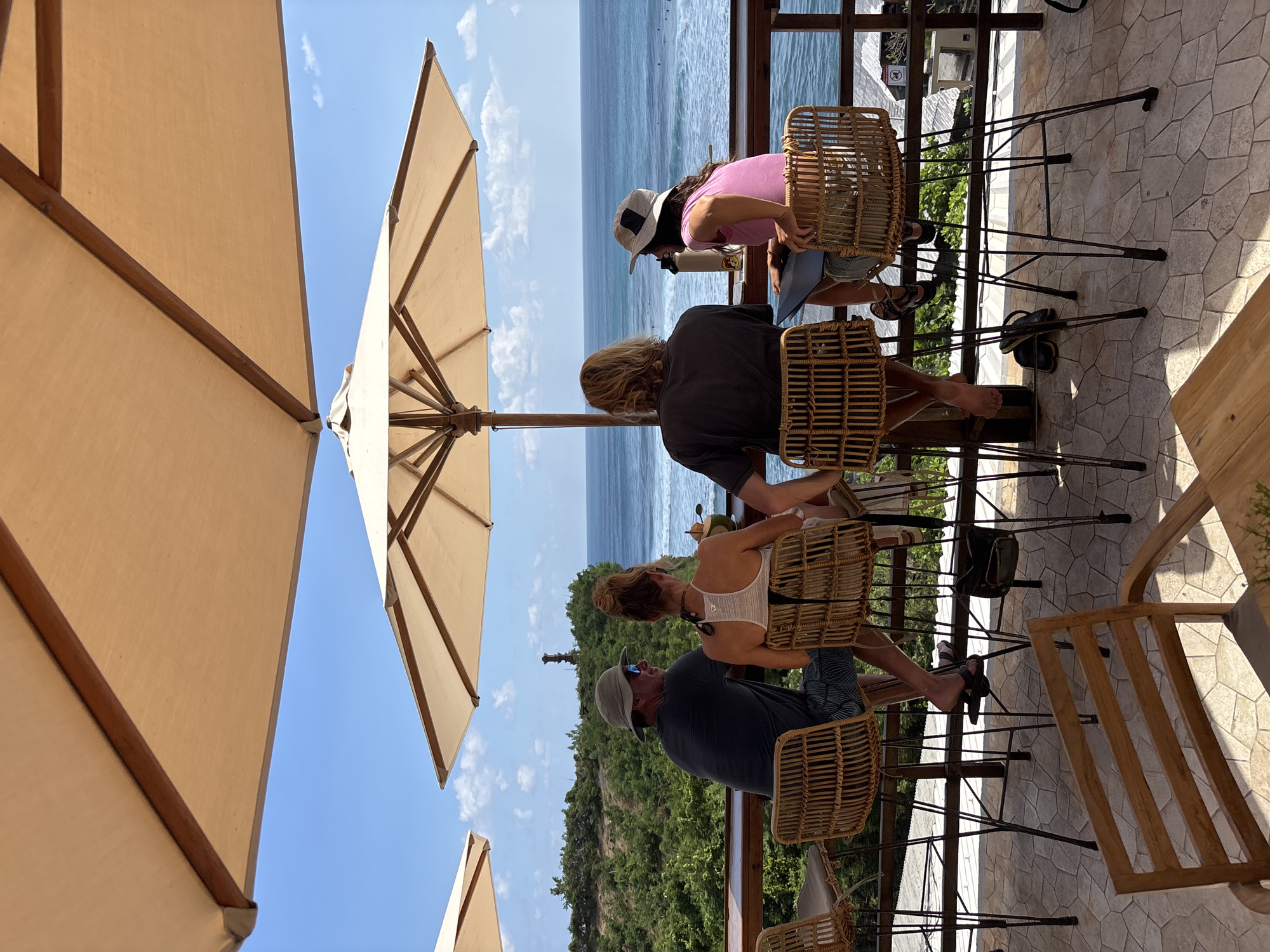 This image depicts an outdoor cafe or restaurant terrace in Kuta Selatan, Indonesia, overlooking the ocean under a clear blue sky with sparse clouds. Four individuals are seated with their backs to the viewer at a high-top wooden counter, facing the sea.
From left to right:
1. A male, wearing a grey t-shirt, patterned shorts, and a light-colored bucket hat with sunglasses. He is looking towards the second person. His left foot, wearing a dark sandal, rests on a lower support. A black purse hangs from the back of his rattan bar stool.
2. A female, wearing a white halter-neck tank top, with her hair tied in a bun. She is seated facing the ocean. A green coconut drink with a straw is on the counter in front of her.
3. A male, with long light-colored hair, wearing a dark t-shirt. He is seated, facing the ocean. His bare feet rest on the stool's footrest, and a pair of dark sandals are placed on the tiled floor beneath his stool.
4. A female, wearing a pink t-shirt and a light-colored bucket hat. She is seated, facing the ocean. A white water bottle and a laptop or tablet are on the counter in front of her. Her feet, wearing sandals, rest on the stool's footrest.
The seating consists of four rattan-style bar stools with black metal legs. Two large, beige square-shaped umbrellas provide shade overhead. A third, smaller umbrella is partially visible in the upper left. The floor is tiled with irregular polygonal light-colored pavers. In the foreground, a partial view of a wooden table and chair suggests additional seating.
Beyond the counter is a wooden railing, followed by a lush green cliffside with dense vegetation. A tall, dark, cylindrical structure (possibly a monument or lighthouse) is visible on the distant cliff between the first and second individuals. The expansive blue ocean dominates the background, with discernible waves and several small figures (surfers) scattered across the water. A light-colored building roof is partially visible below the railing on the far right. The scene is illuminated by bright daylight, casting distinct shadows.
