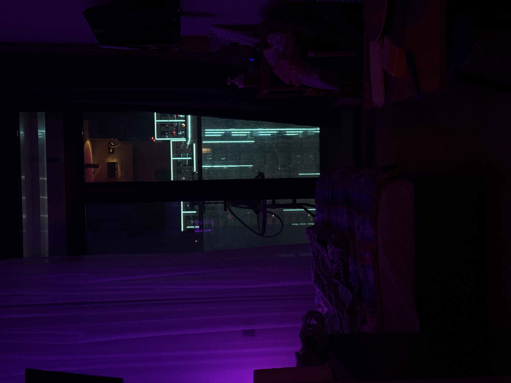 The image is an interior shot taken at night, with a strong purple hue dominating the scene. The location appears to be a room with a large window offering a view of a city skyline. The skyline visible through the window suggests a dense urban environment, with brightly lit buildings in the background. The window is large and wide, giving a panoramic view of the city.
Inside the room, there is a fabric-draped chair or couch in the foreground to the right. The fabric has a patterned design with dark colors, adding texture and contrast to the scene. The purple ambient lighting casts a surreal glow on the room's surfaces, making it hard to discern specific details in the shadows.
The image is turned upside-down. Considering the architectural style of the visible buildings and the general environment, it's unlikely to be Sochi, Russia, as it does not resemble known architectural features of that city. Without further context, the exact location cannot be verified.
