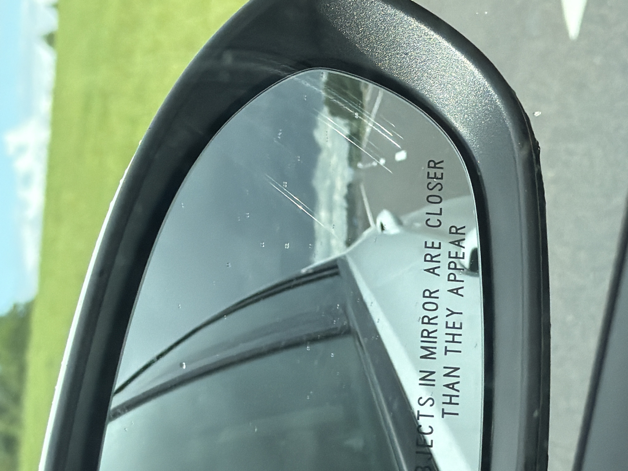 The image is a close-up, angled shot of a car's side-view mirror. The mirror is reflecting a cloudy sky, a road, and a grassy field with trees in the distance. The text "OBJECTS IN MIRROR ARE CLOSER THAN THEY APPEAR" is visible on the lower portion of the mirror. There are several water droplets on the mirror's surface and some faint scratches. The exterior of the mirror housing is black and textured. The scene appears to be outdoors, possibly on a highway, given the road reflection. The location is stated to be in the Пяршайскі сельскі Савет, Belarus, though this context is not directly discernible from the visual elements of the image itself. There are no people or explicit actions depicted in the image.