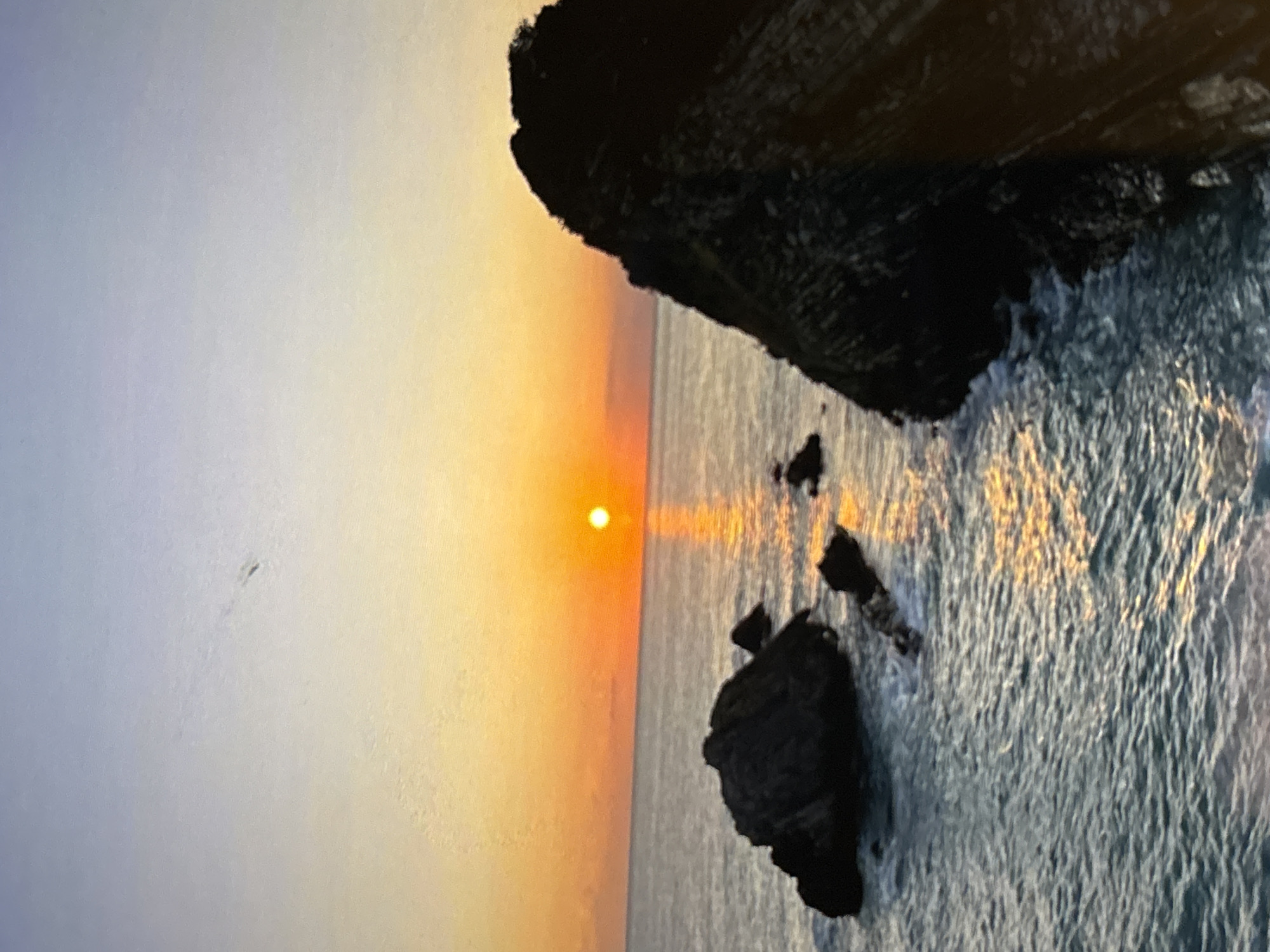 The image depicts a coastal sunset. A bright sun is positioned low in the sky, casting a golden reflection on the ocean. Silhouetted rock formations are visible along the coastline. There are no people visible. The location is not identifiable as Belopoptsi, Bulgaria, from visual details alone.