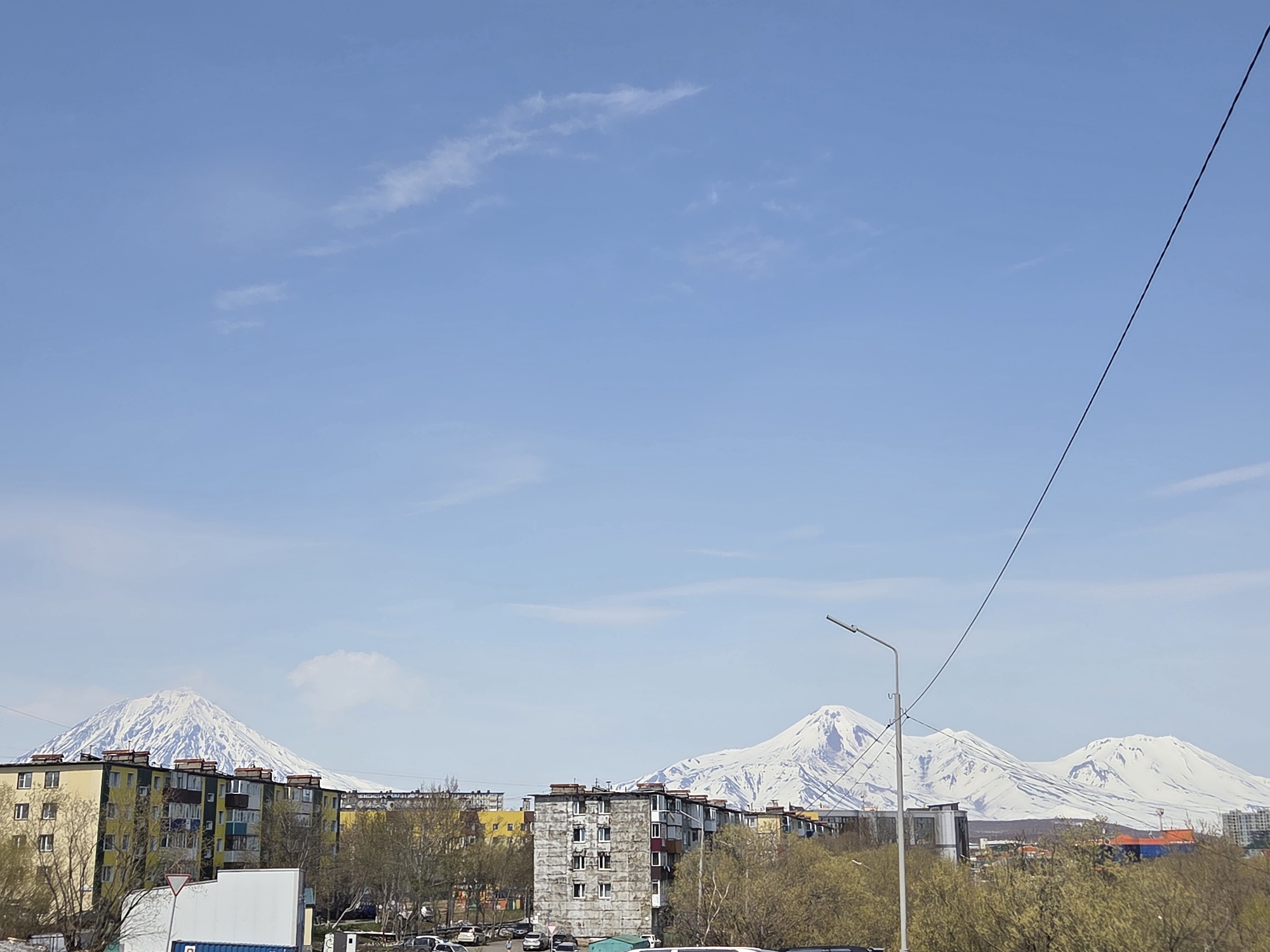 The image depicts a cityscape with a clear blue sky punctuated by scattered clouds. In the background, a mountain range capped with snow dominates the horizon. Closer to the foreground are several residential buildings of varying colors and conditions. Some of the buildings appear to be apartment complexes. Trees with budding leaves are interspersed among the buildings. A utility pole and overhead wires are visible on the right side of the frame. The ground level shows cars parked along the streets. The location appears to be a city, but its specific name is unknown.
