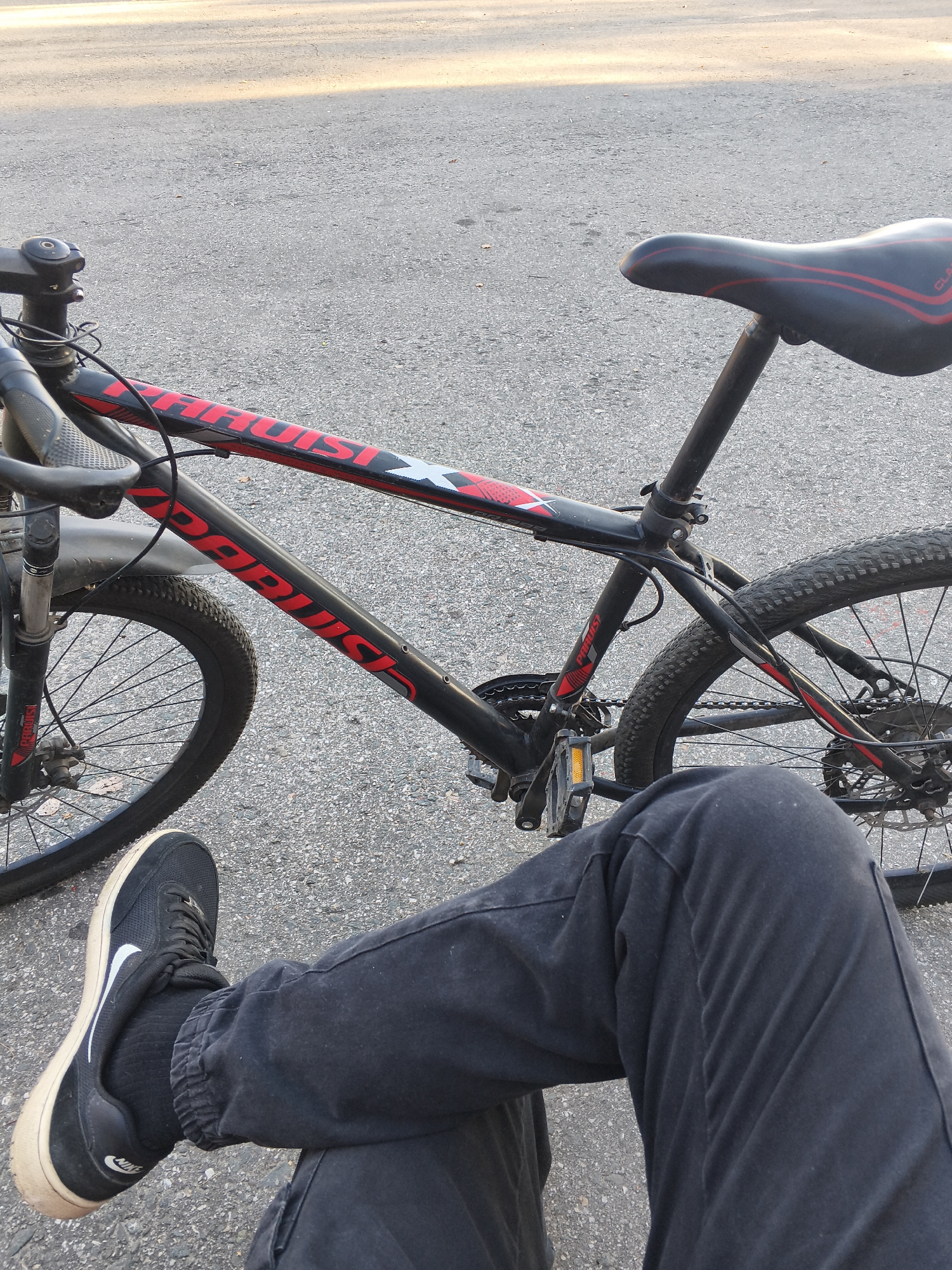 A person's legs, clad in black pants and black Nike sneakers, are positioned in the foreground, obscuring a portion of a black bicycle with red accents. The bicycle is parked on an asphalt surface, likely a street or pathway, as indicated by the textured grey ground. Sunlight casts shadows and highlights on the scene, suggesting it is daytime. The bicycle features a red seat with black trim and red lettering that reads "THRUST" on the frame. The front wheel and part of the front fork are visible to the left, while the rear wheel and drivetrain are to the right. The overall impression is a casual, outdoor setting. The location of Mokshan, Russia, is provided as context.