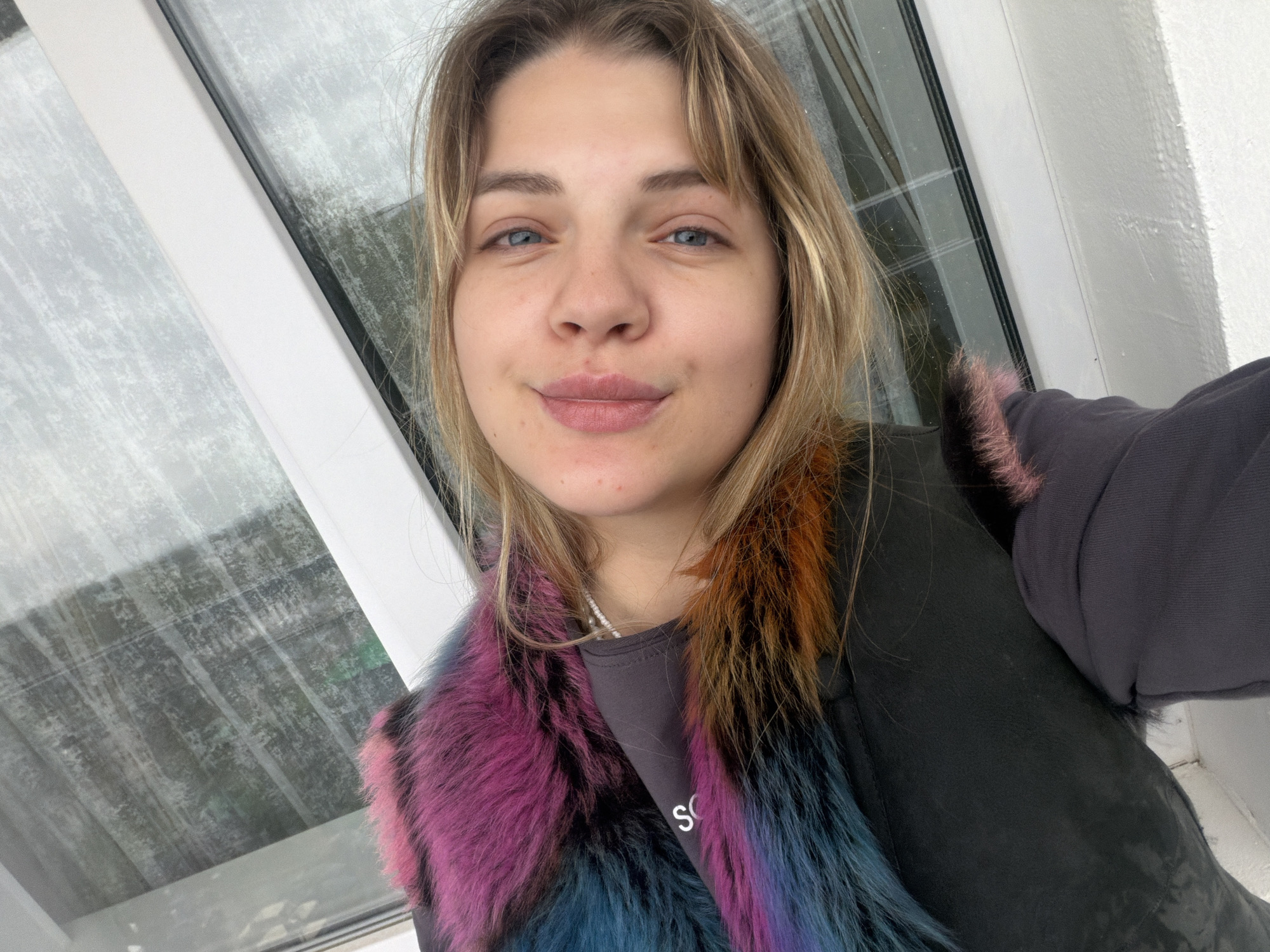 A young woman is taking a selfie indoors. She has blonde hair with bangs, blue eyes, and fair skin. She is wearing a gray top and a black vest with colorful faux fur trim around the collar, featuring shades of purple, orange, and blue. The background shows a window with a blurry view of what appears to be a snowy or rainy scene. The location might be in the city of Chaikovsky, Russia, but this cannot be definitively confirmed from the image alone.