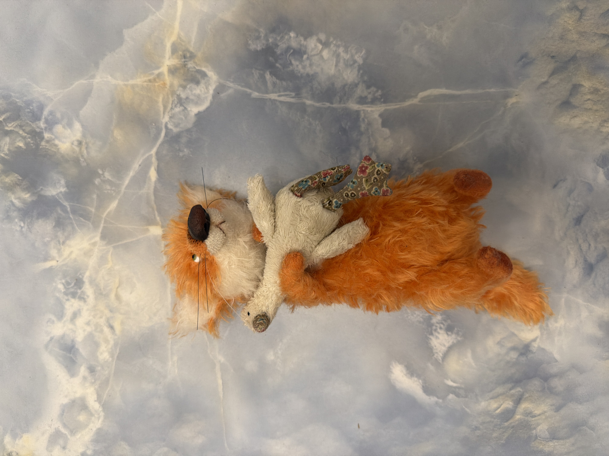 A plush toy fox with orange fur and a white chest is positioned vertically in the center of the frame, clutching a small, white plush bird. The fox's eyes are green, and it has a dark brown nose and whiskers. The bird has a white body and a small, dark beak. The background is a marble-like surface with blue, white, and beige marbled patterns.
