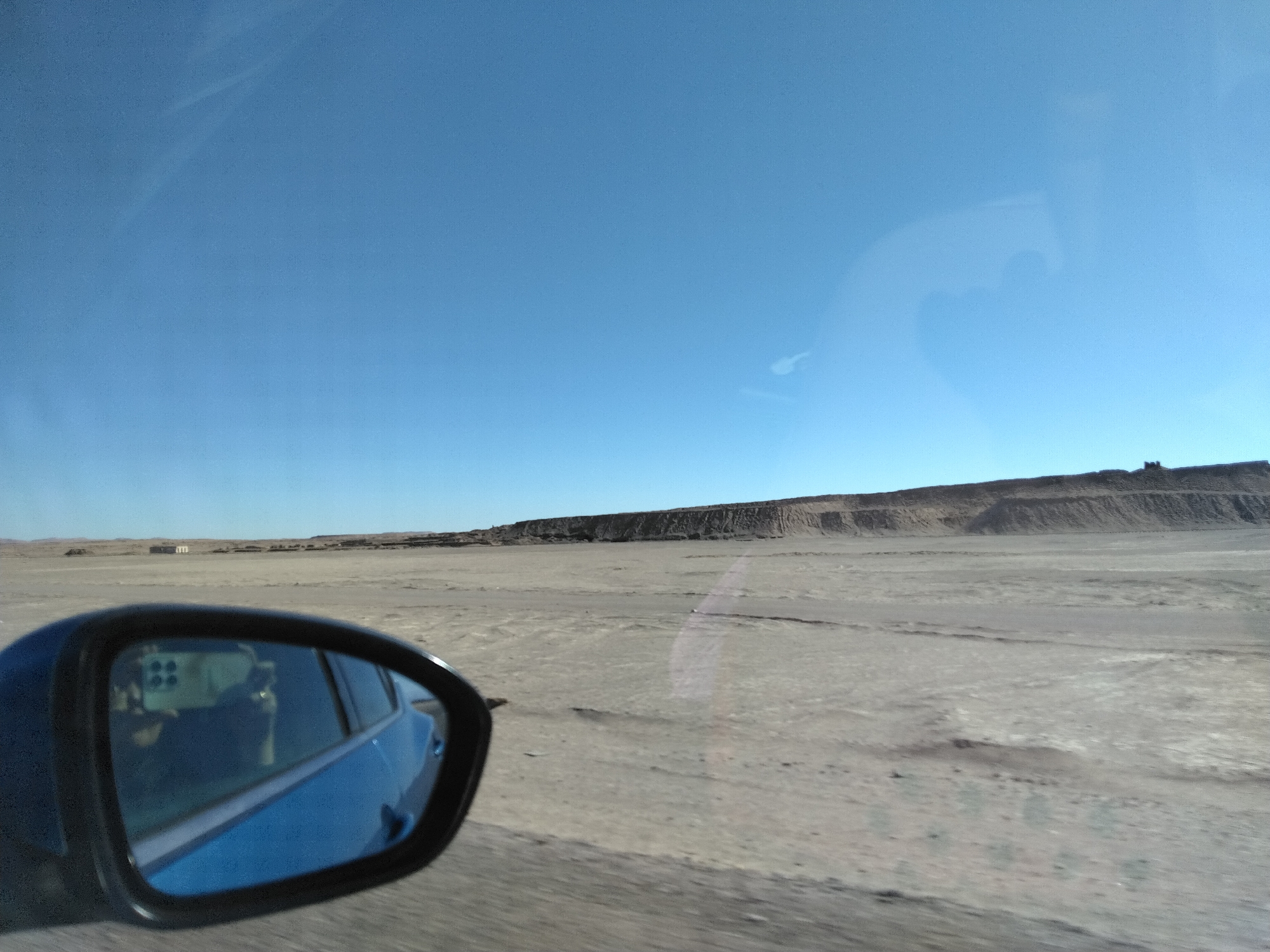 The media file shows a view from inside a car, looking out towards a vast, dry, and barren landscape under a clear blue sky. A side-view mirror in the foreground reflects the interior of the car, including what appears to be passengers and possibly a camera or phone mounted on the dashboard. The landscape itself is mostly flat, sandy, and appears arid, with a distant low ridge or cliff line forming the horizon. There are a few structures visible in the far distance, possibly buildings or industrial equipment, suggesting a remote, possibly desert or steppe environment. The time of day appears to be daytime, with bright sunlight casting minimal shadows, indicating the sun is high in the sky. The weather seems clear and dry. The scene evokes a sense of travel through a remote and undeveloped area, with a feeling of openness and isolation. No text is visible in the image.